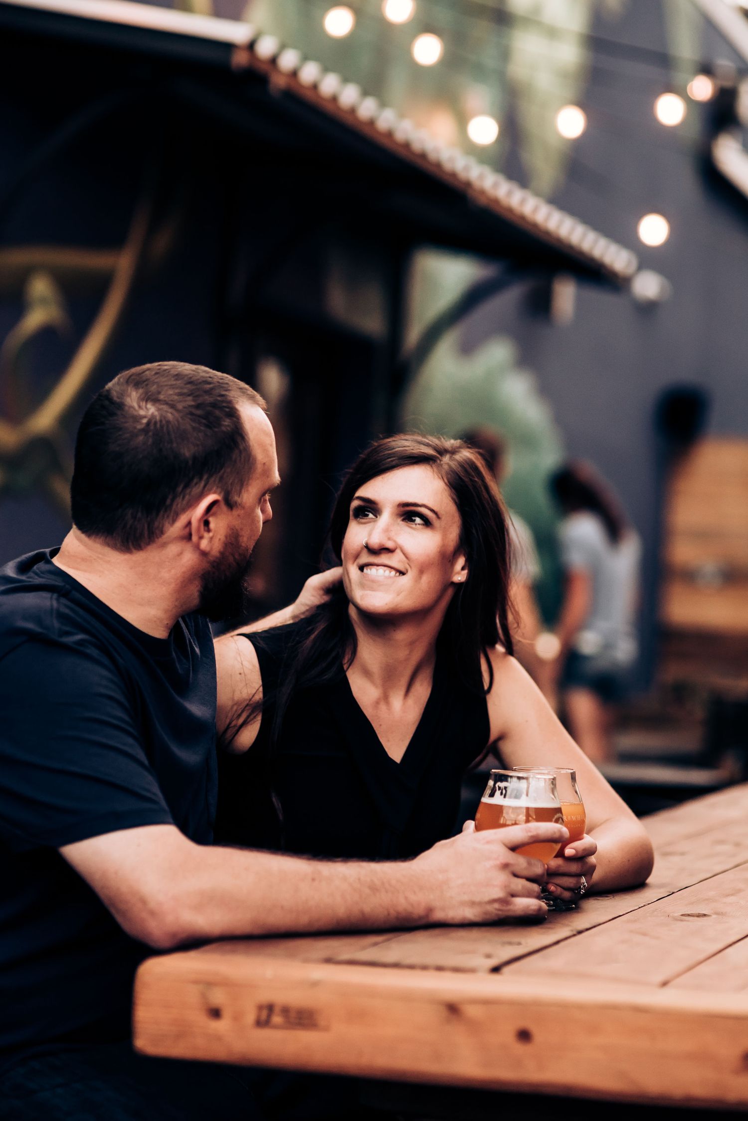 Couple sharing laughter over craft beer at rustic outdoor table.