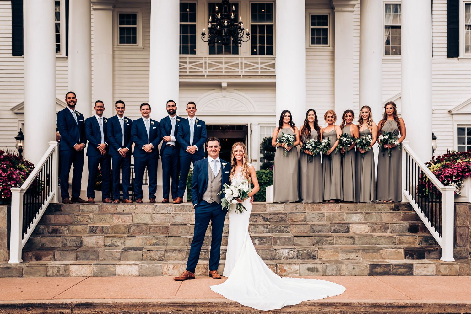 Wedding party poses on grand white mansion steps with floral decorations.