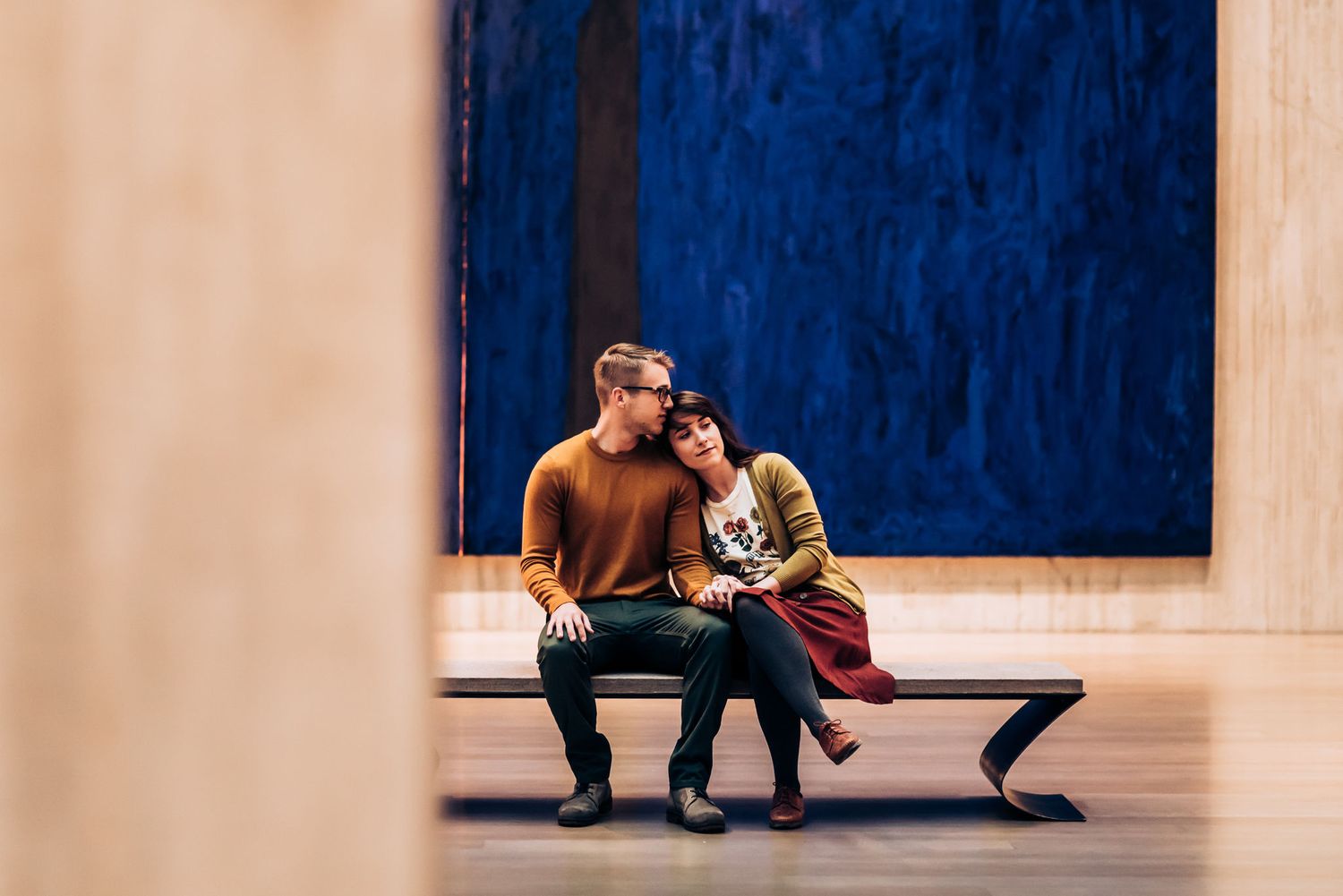 A couple sits intimately on a bench in an art gallery with a large blue artwork displayed behind them.