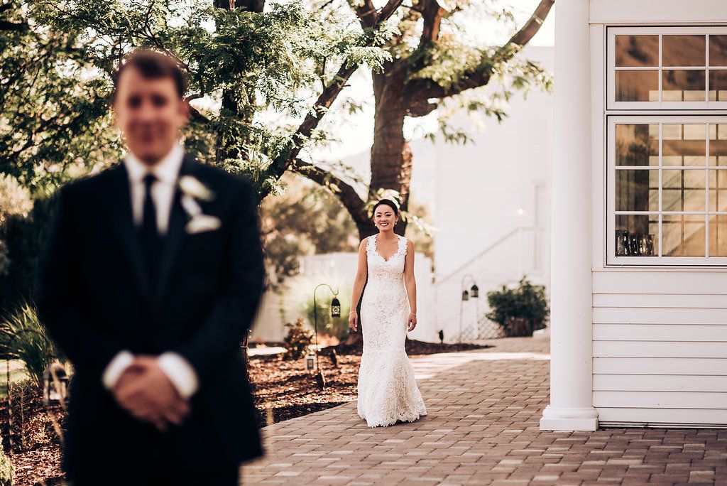 Wedding first look moment at elegant white venue with bride in distance and groom in foreground.
