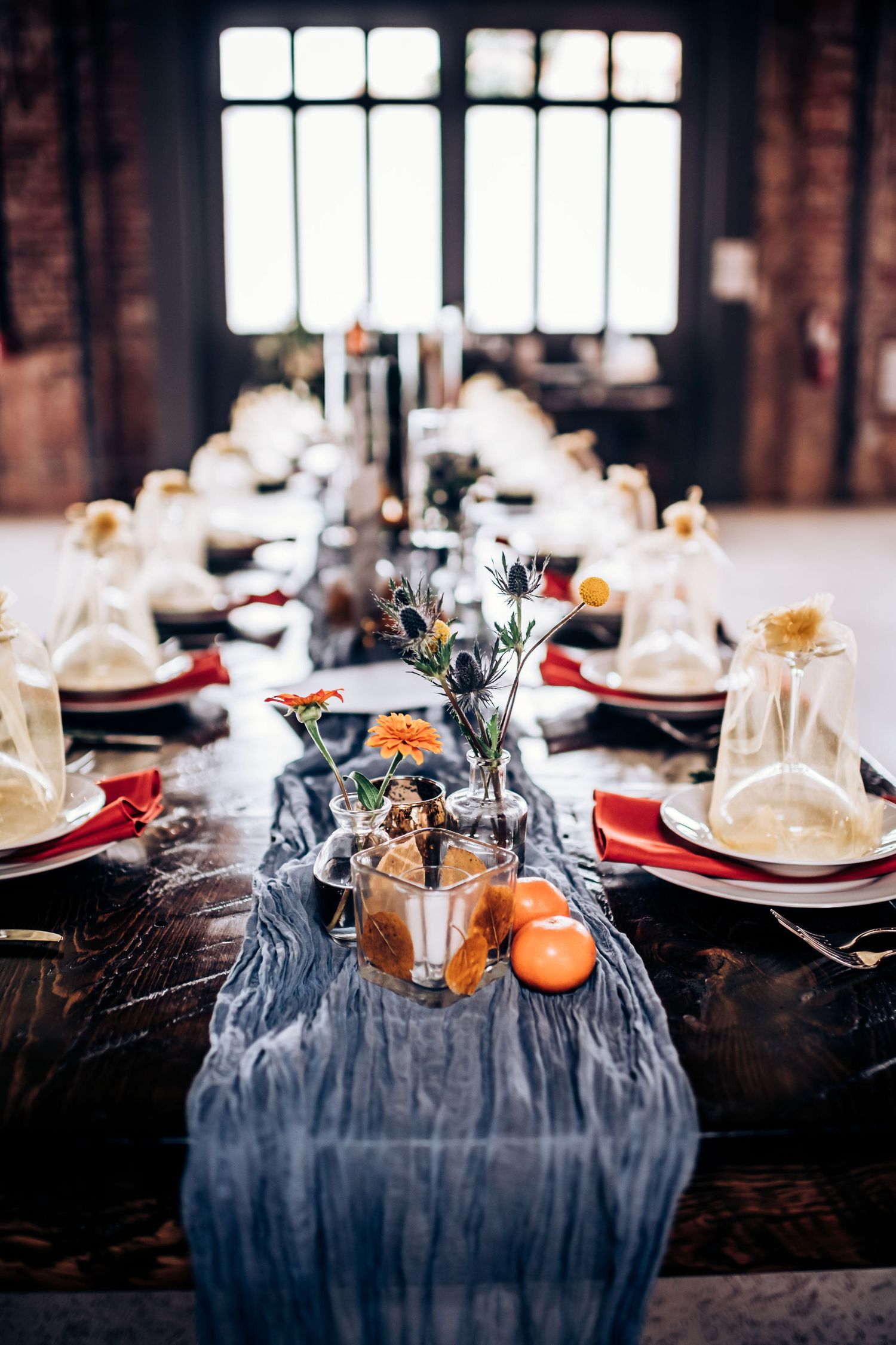 Elegant table setting with navy runner, oranges, and white plates in rustic venue with large windows.