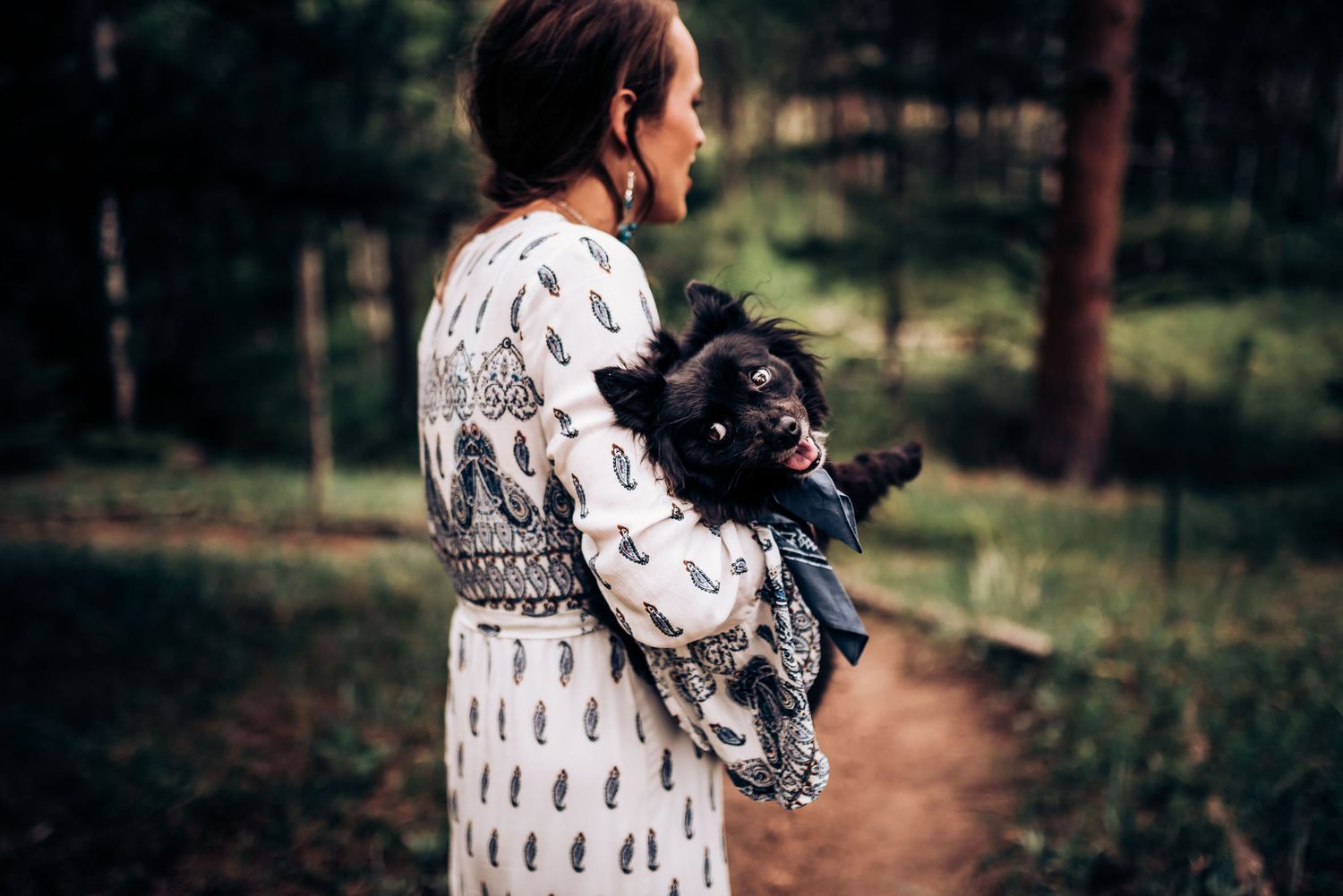 Person wearing a patterned white dress holds a black dog in a wooded setting.