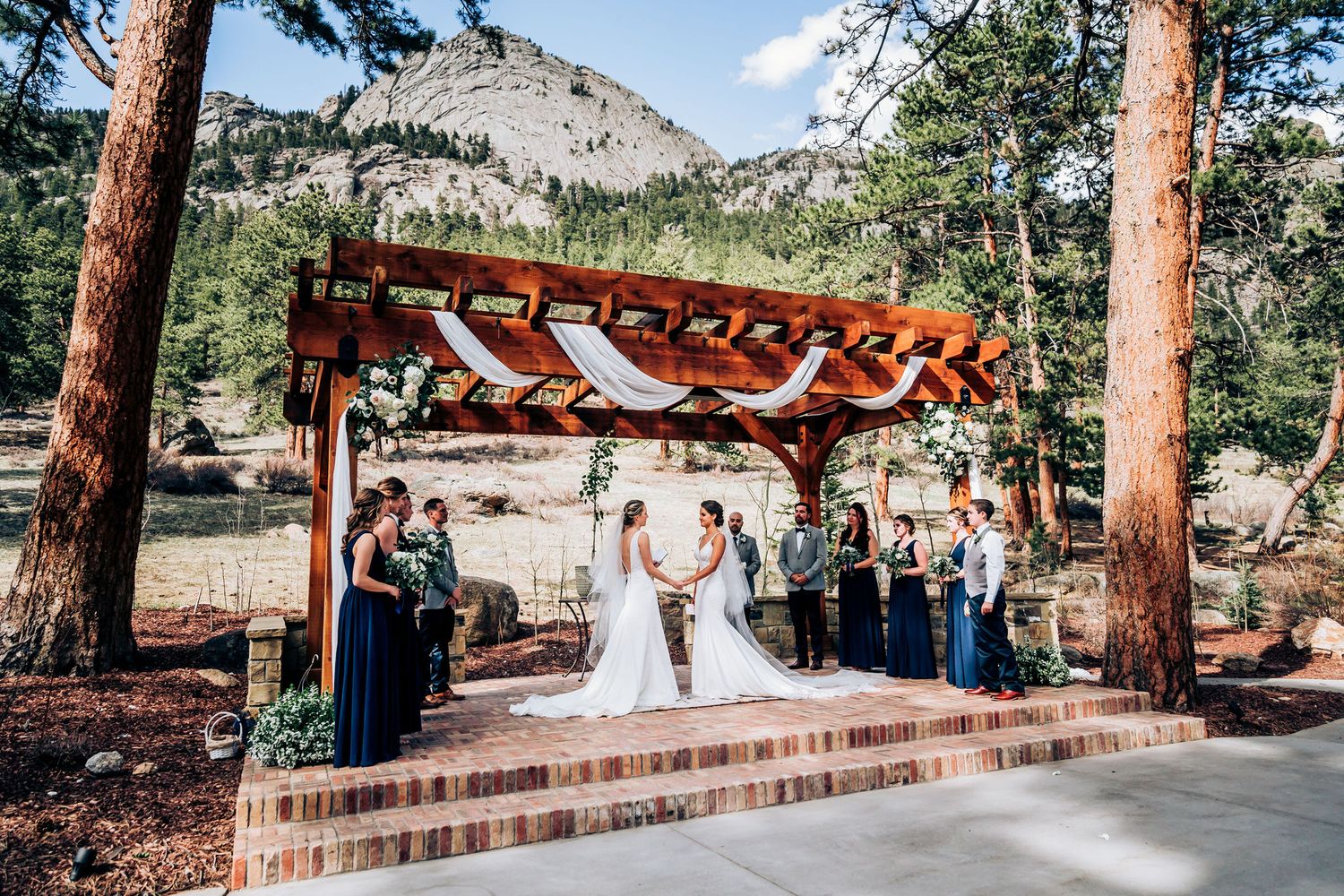 Mountain wedding ceremony under wooden pergola with dramatic rocky backdrop.