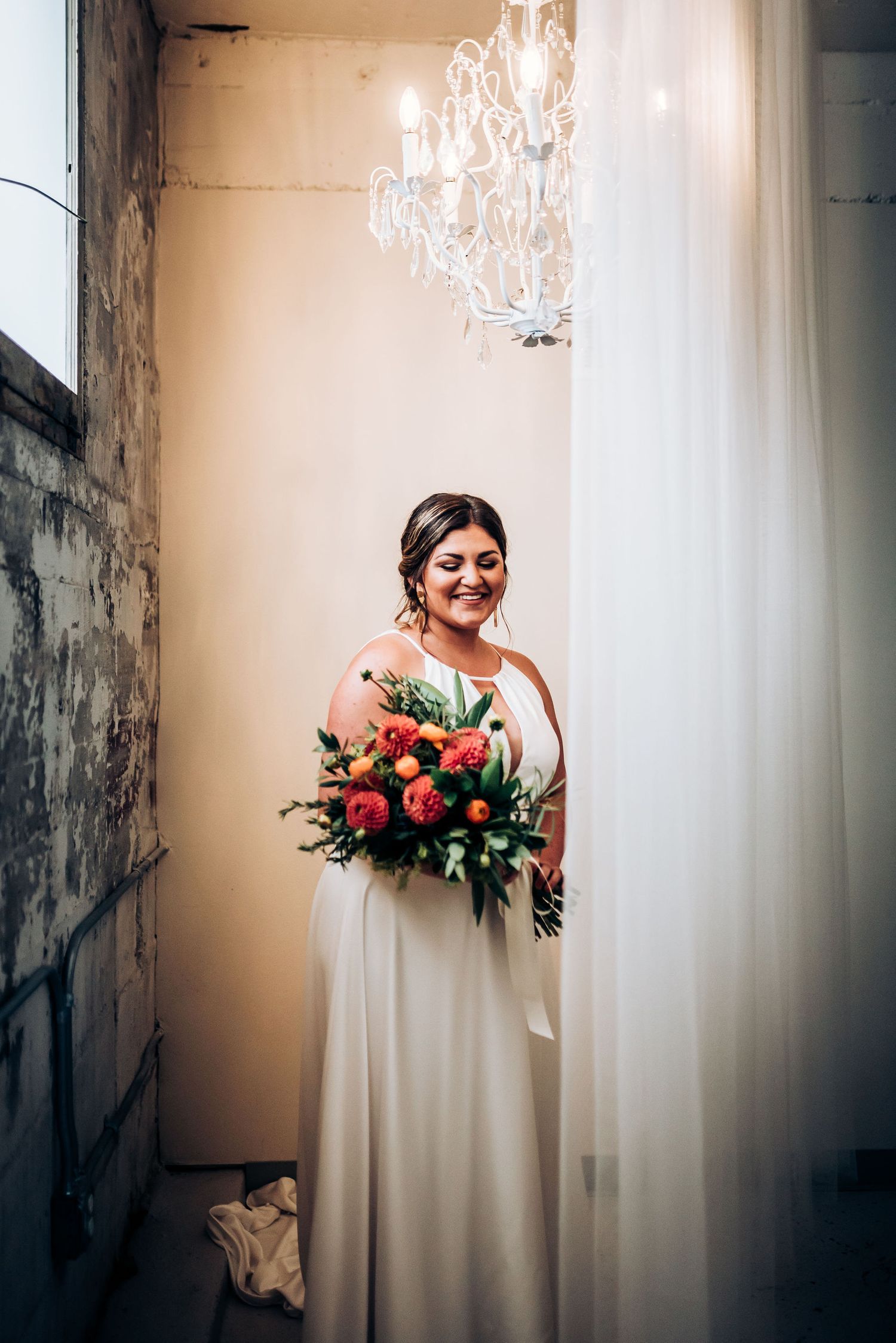 Bride in elegant white gown holds colorful bouquet beneath crystal chandelier.