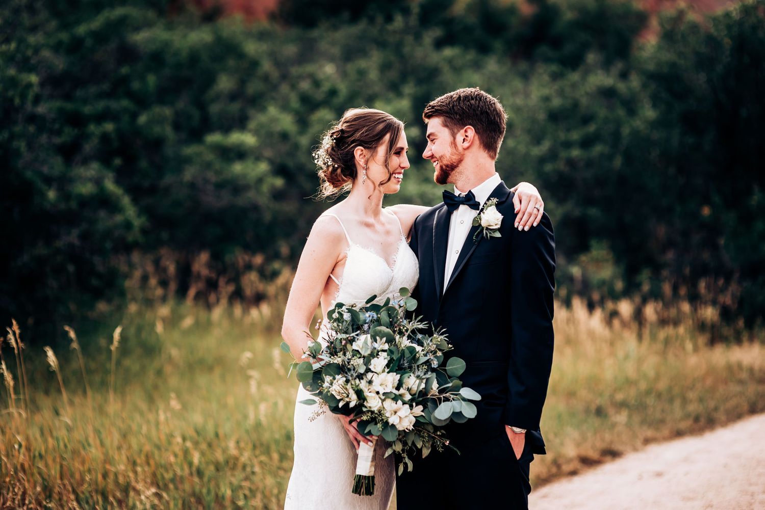 Elegant wedding couple posing in golden field with bouquet of white and green flowers.