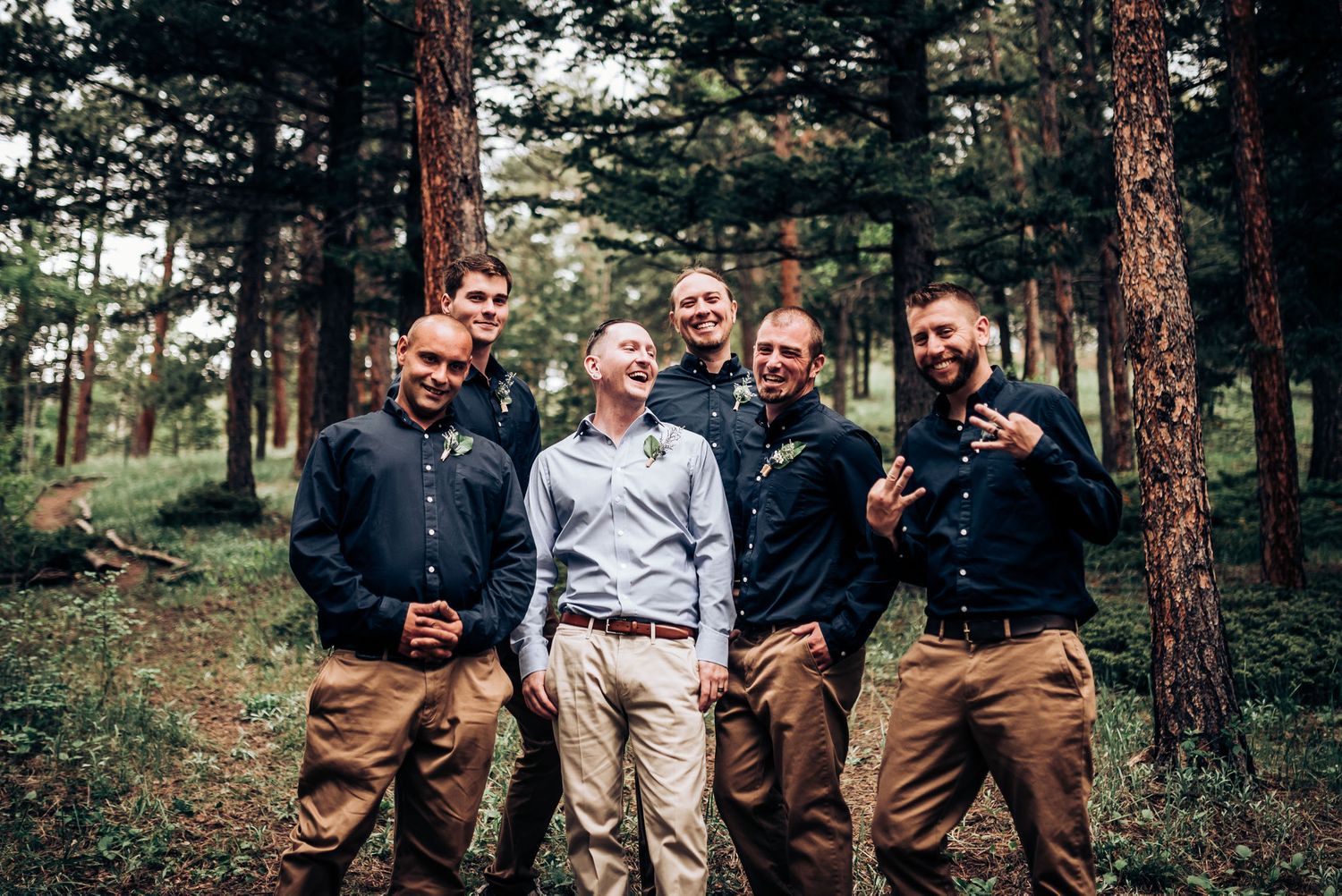 Groomsmen in coordinated attire pose casually in pine forest.
