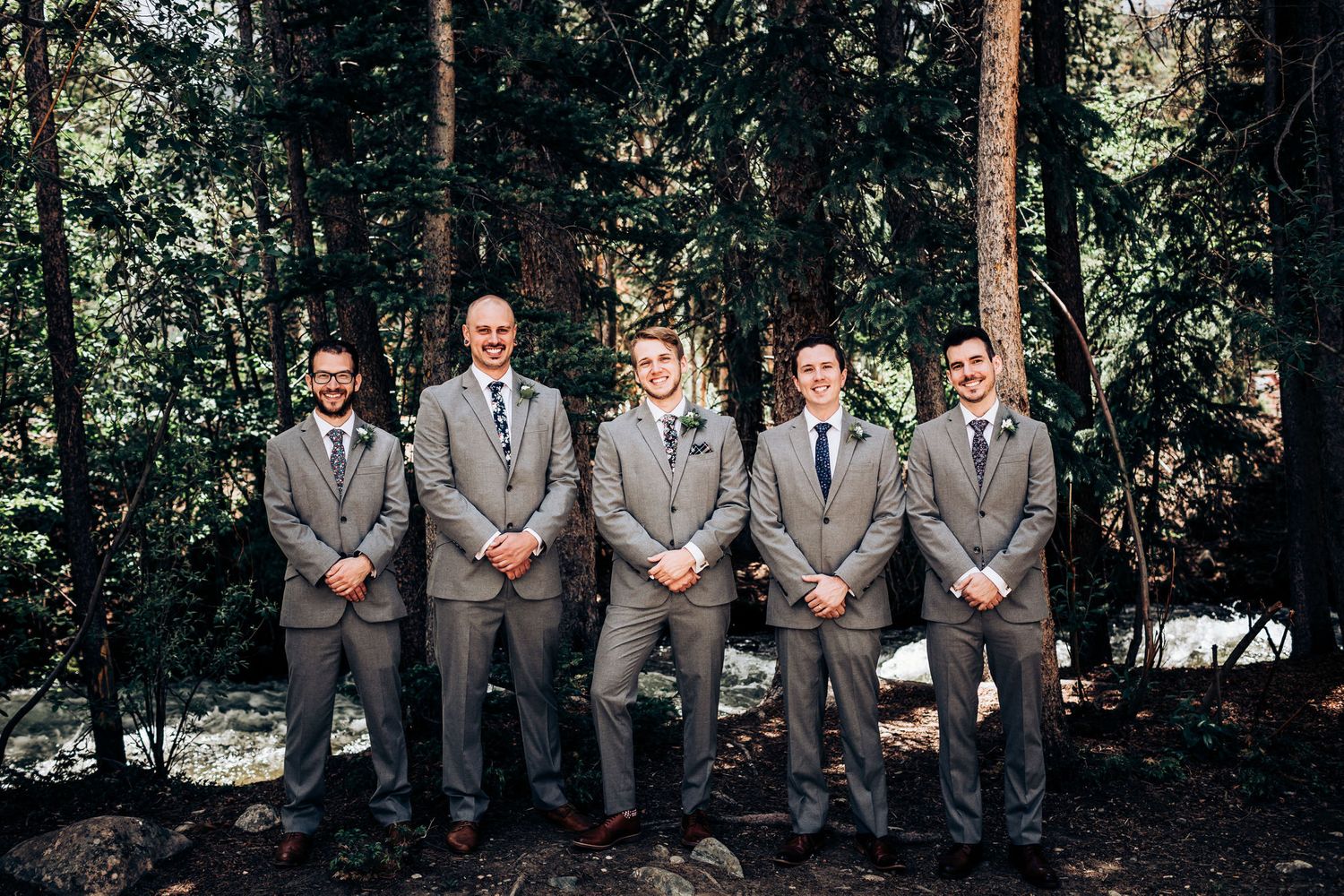 Groomsmen in matching gray suits pose formally against forest backdrop.