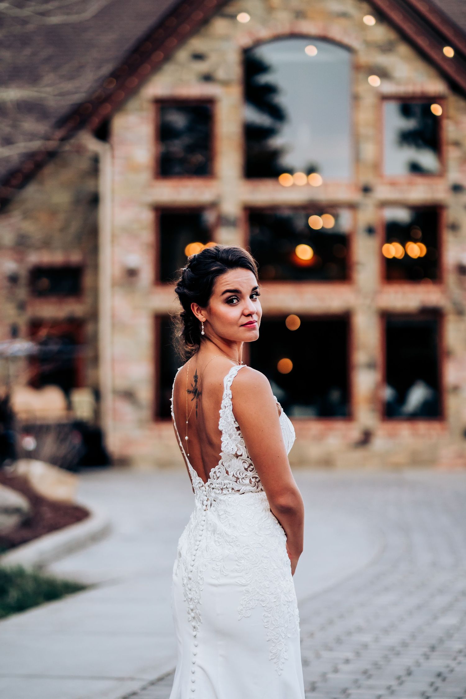 An elegant wedding dress with lace detailing and open back design photographed against a stone building backdrop.