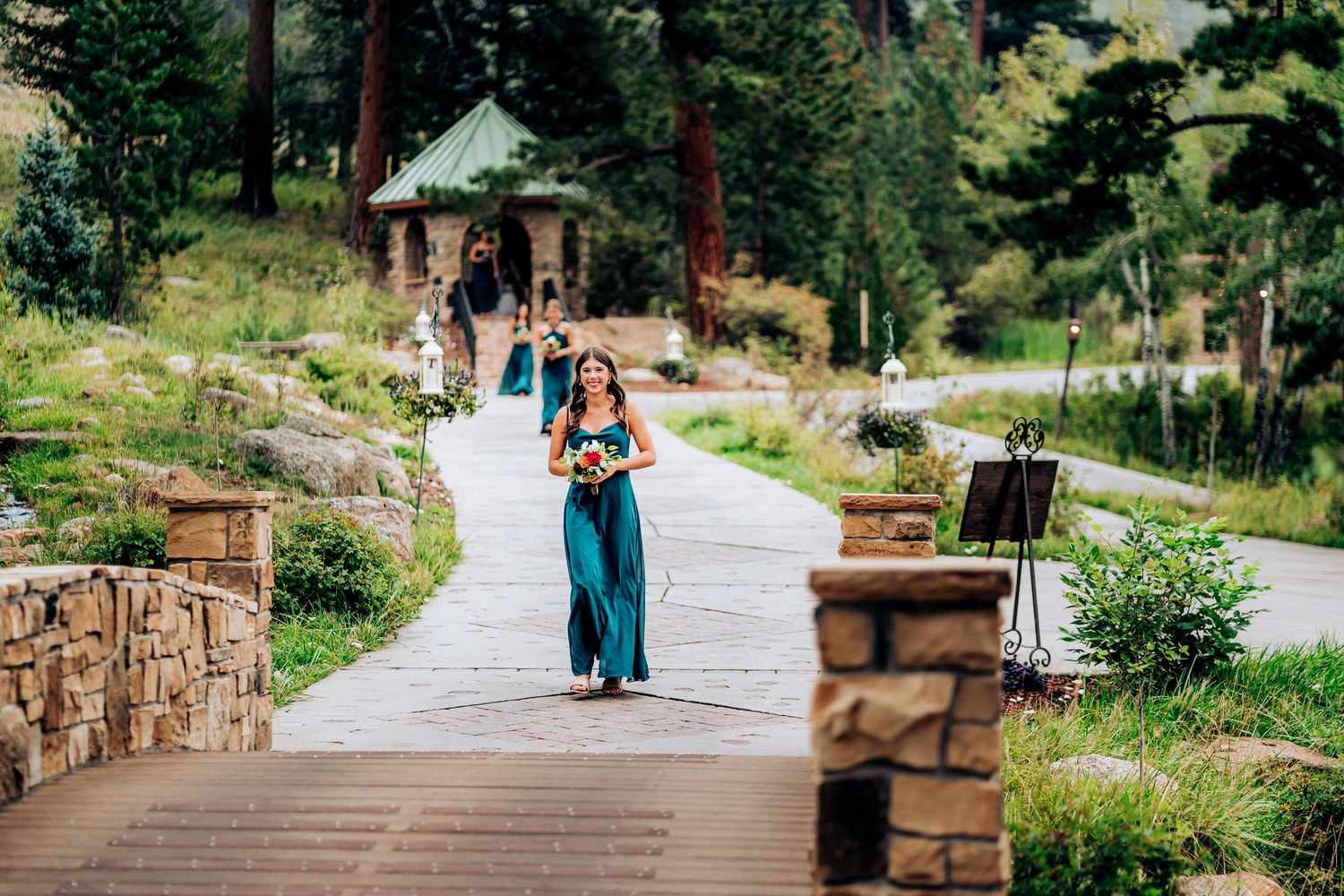 Wedding attendant in teal dress walking down stone-lined path with gazebo in background.