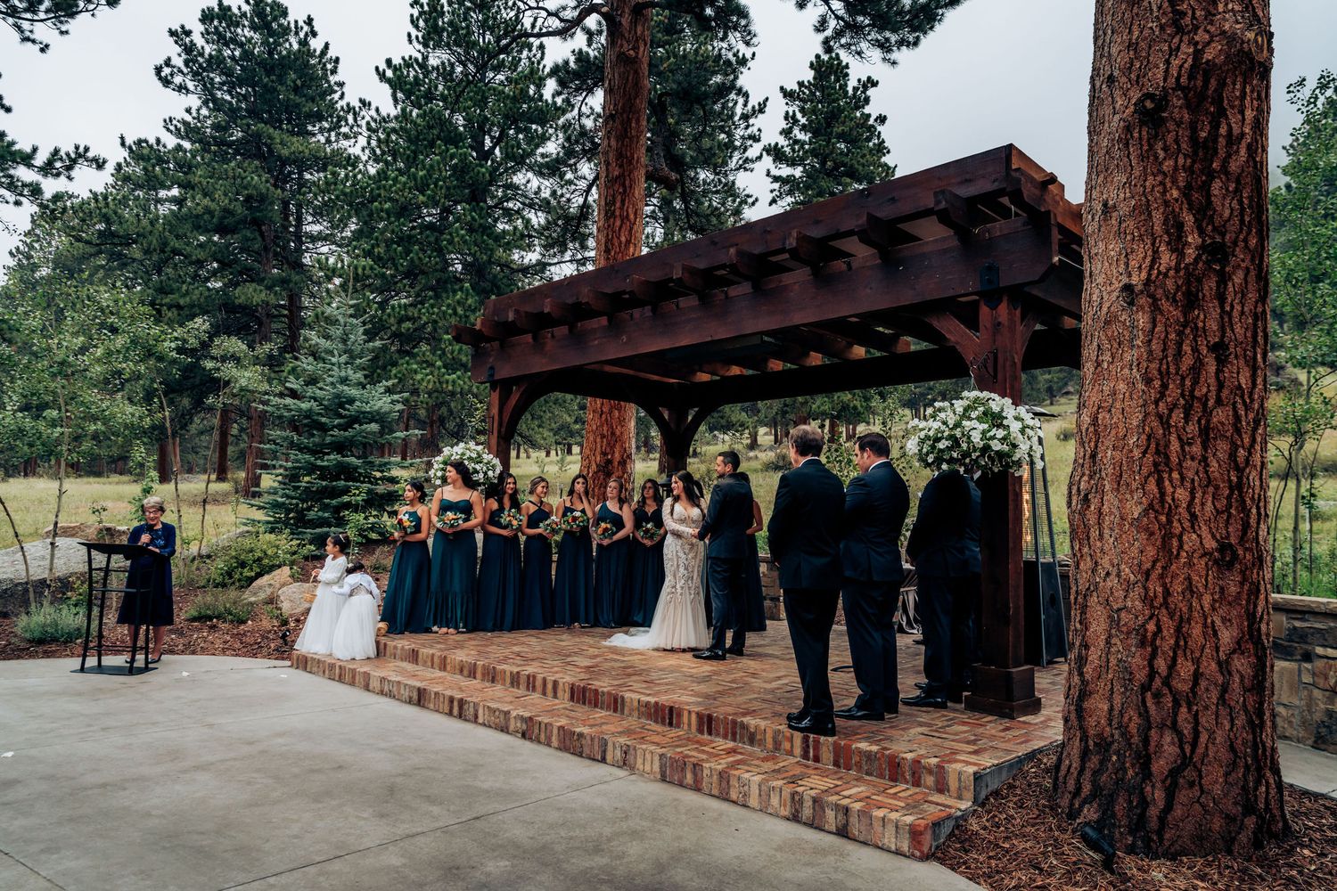 Outdoor wedding ceremony under wooden pergola surrounded by tall pine trees.