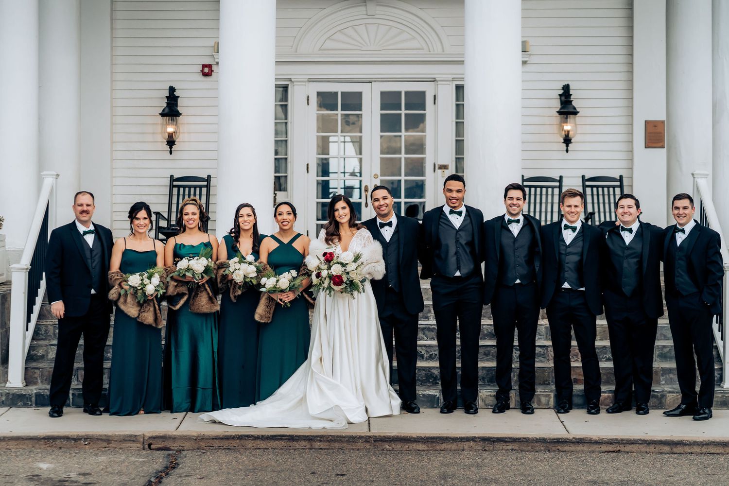 Wedding party lined up on white porch with emerald green bridesmaid dresses and black tuxedos.