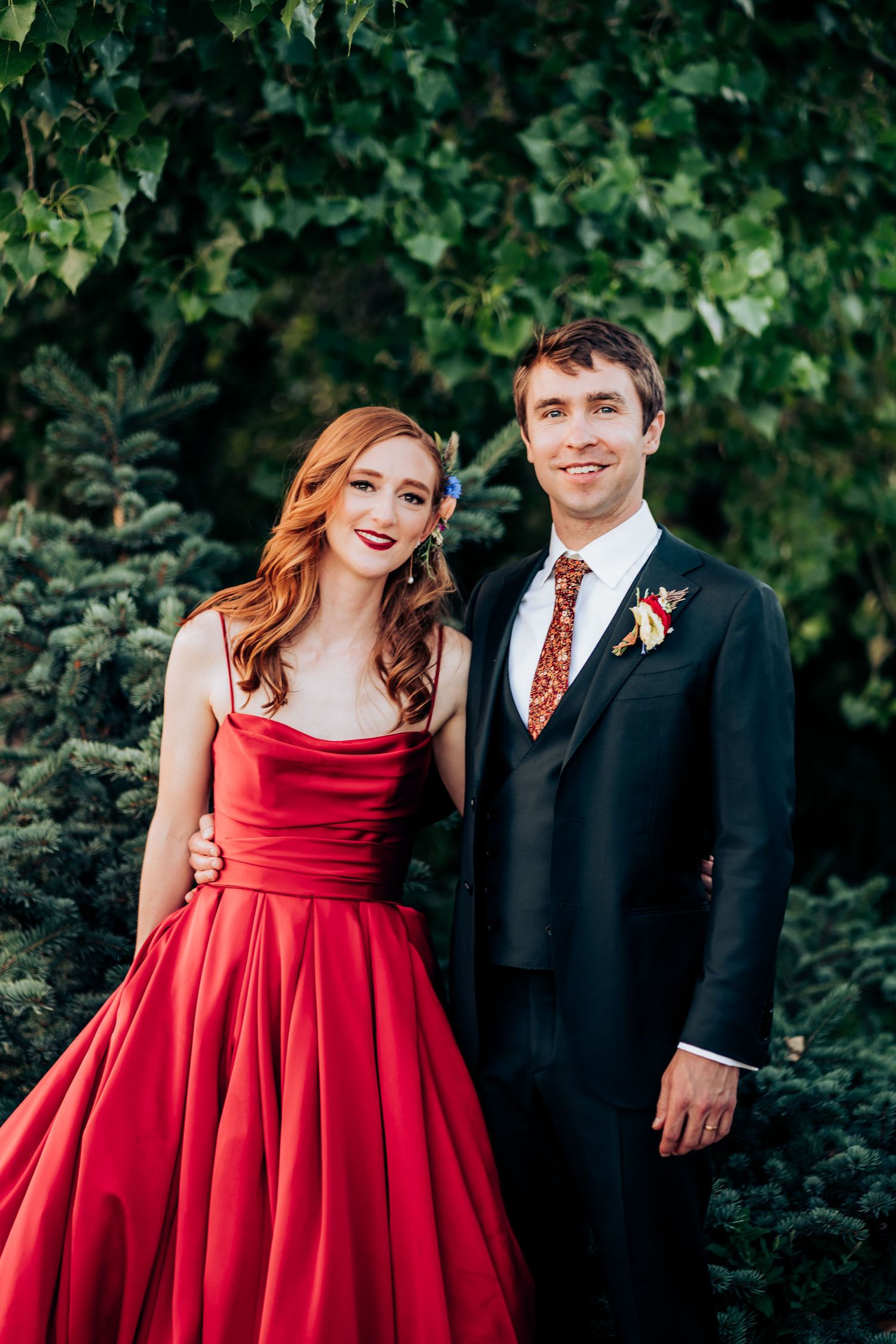 Bride in stunning red satin ballgown poses with groom against lush garden backdrop.