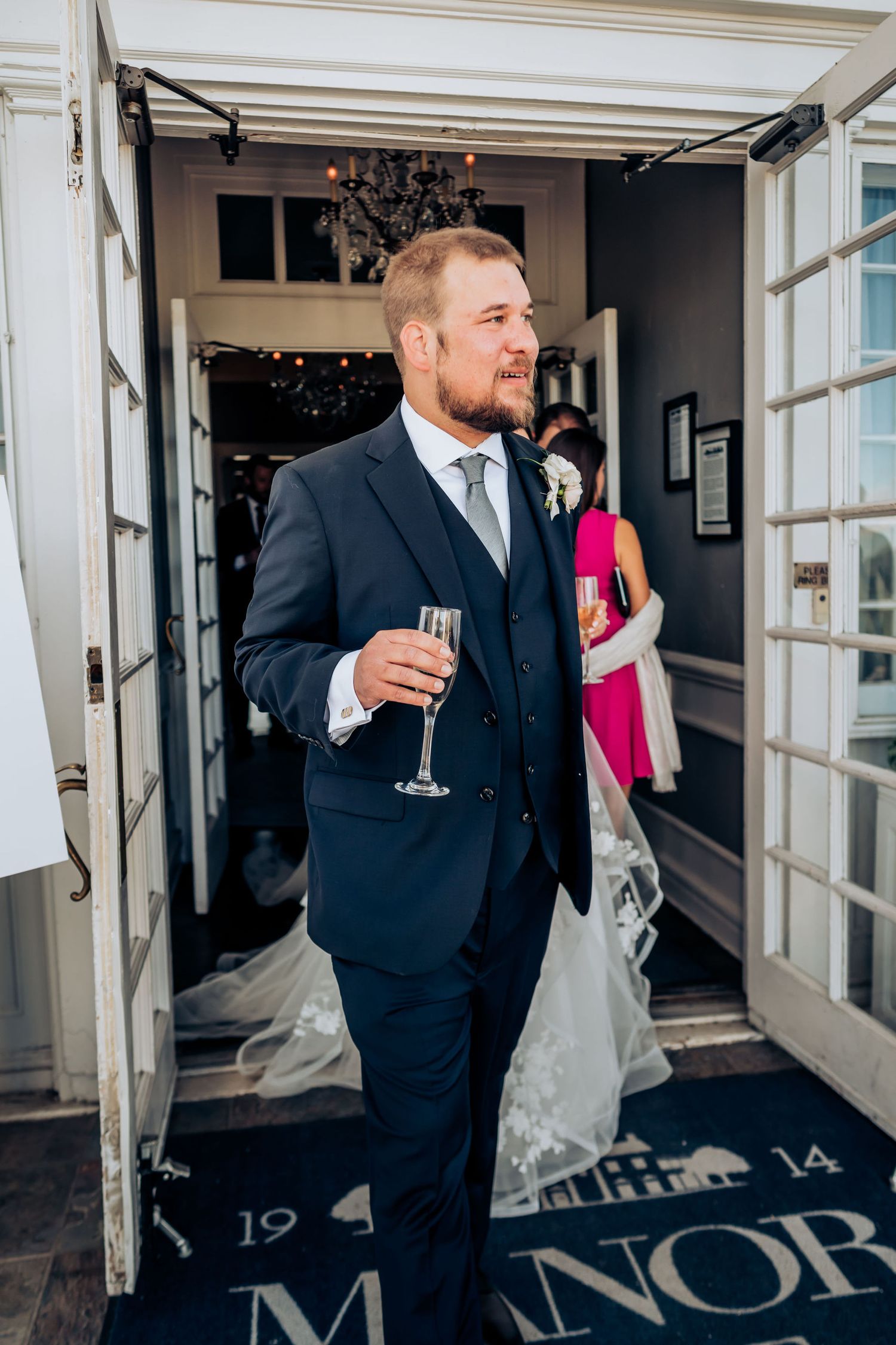Groom holding champagne glass in doorway of manor house during wedding reception.