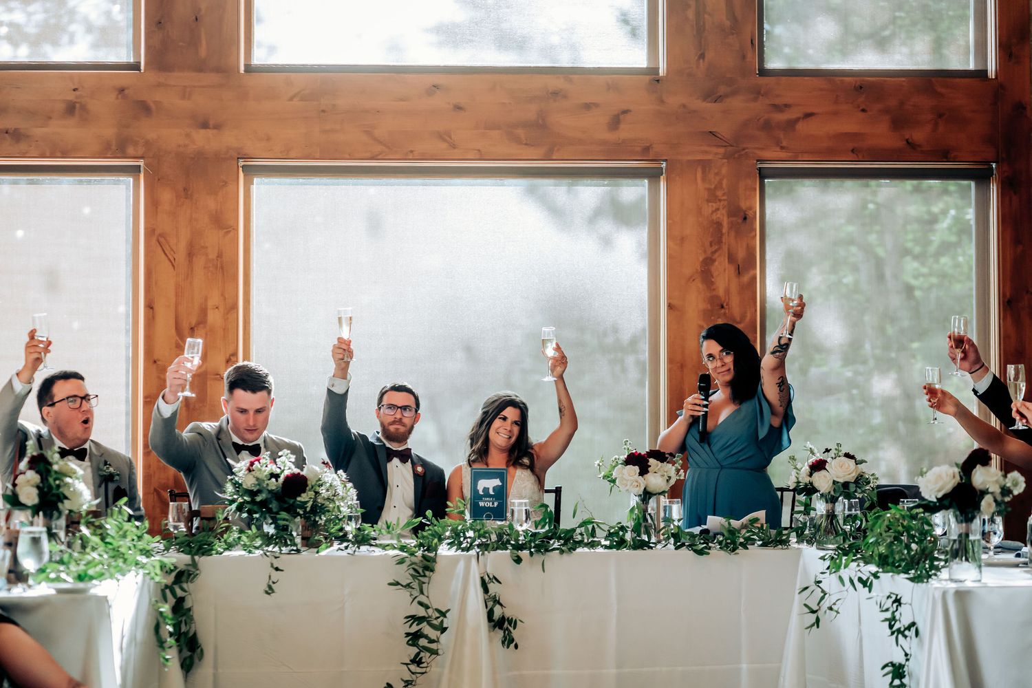 Wedding party members raise their glasses in celebration at a head table decorated with greenery.