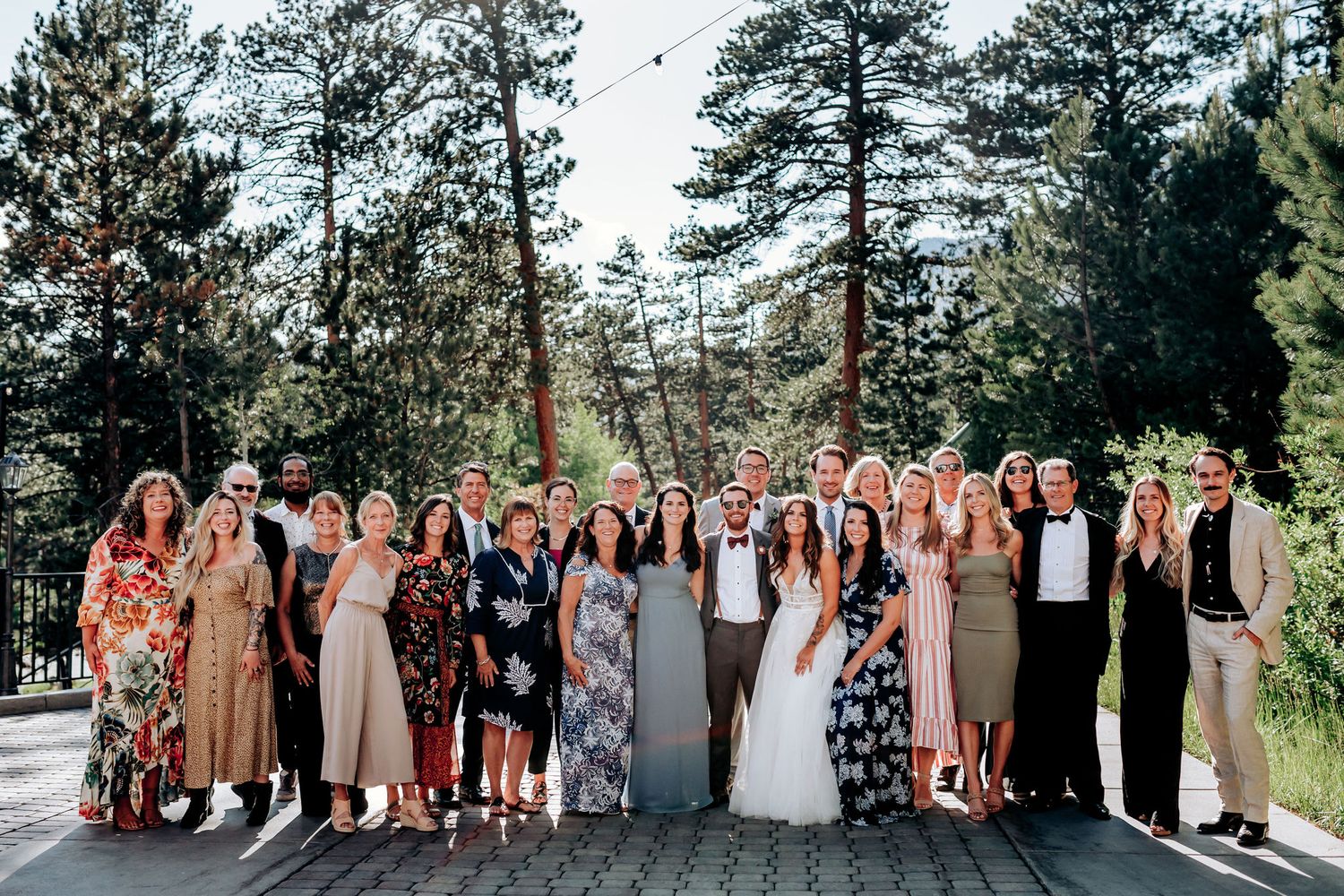 Large wedding group photo with guests in colorful attire against mountain forest backdrop.