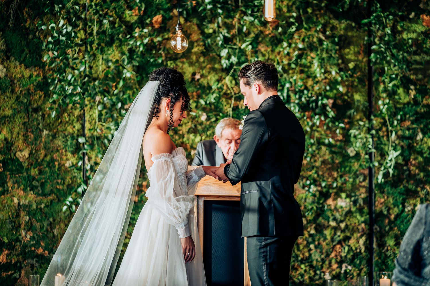 Intimate outdoor wedding ceremony with verdant wall backdrop and suspended lanterns.