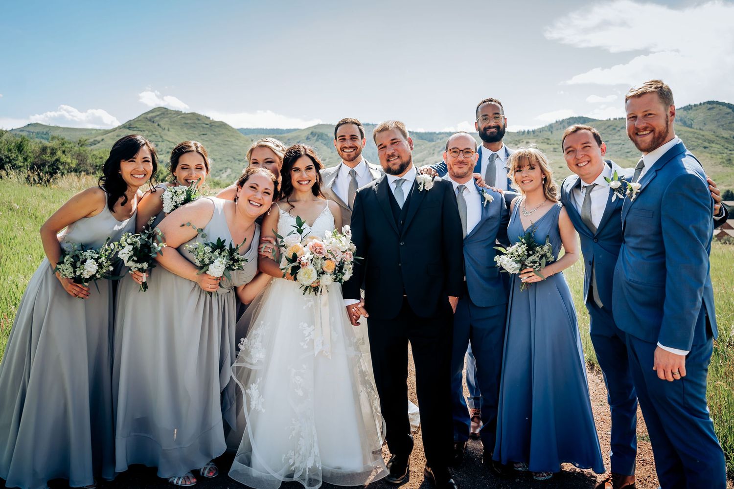 Wedding party in blue and gray attire posing for formal photo in mountain field.