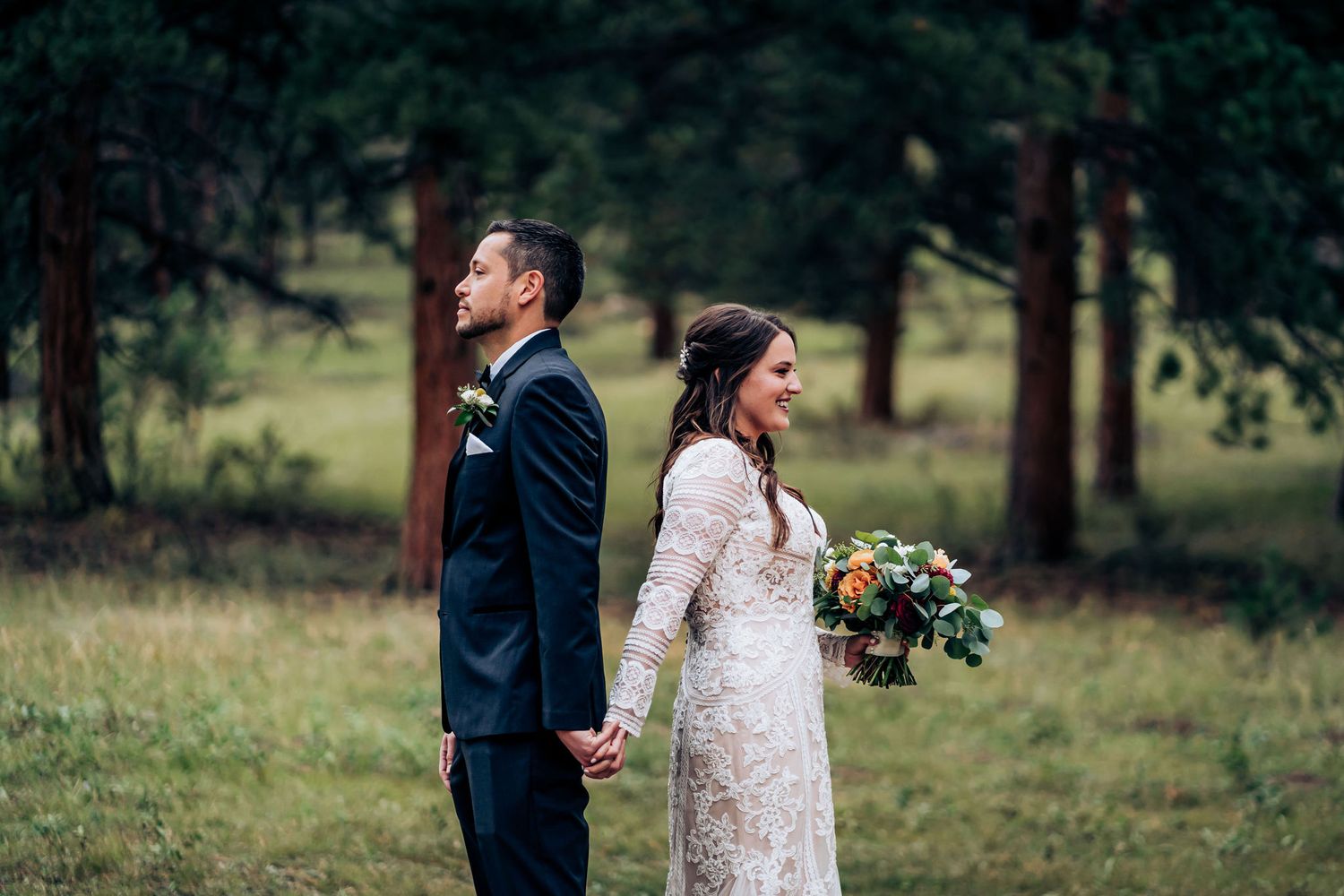 Wedding couple stands back-to-back in forest clearing holding hands.