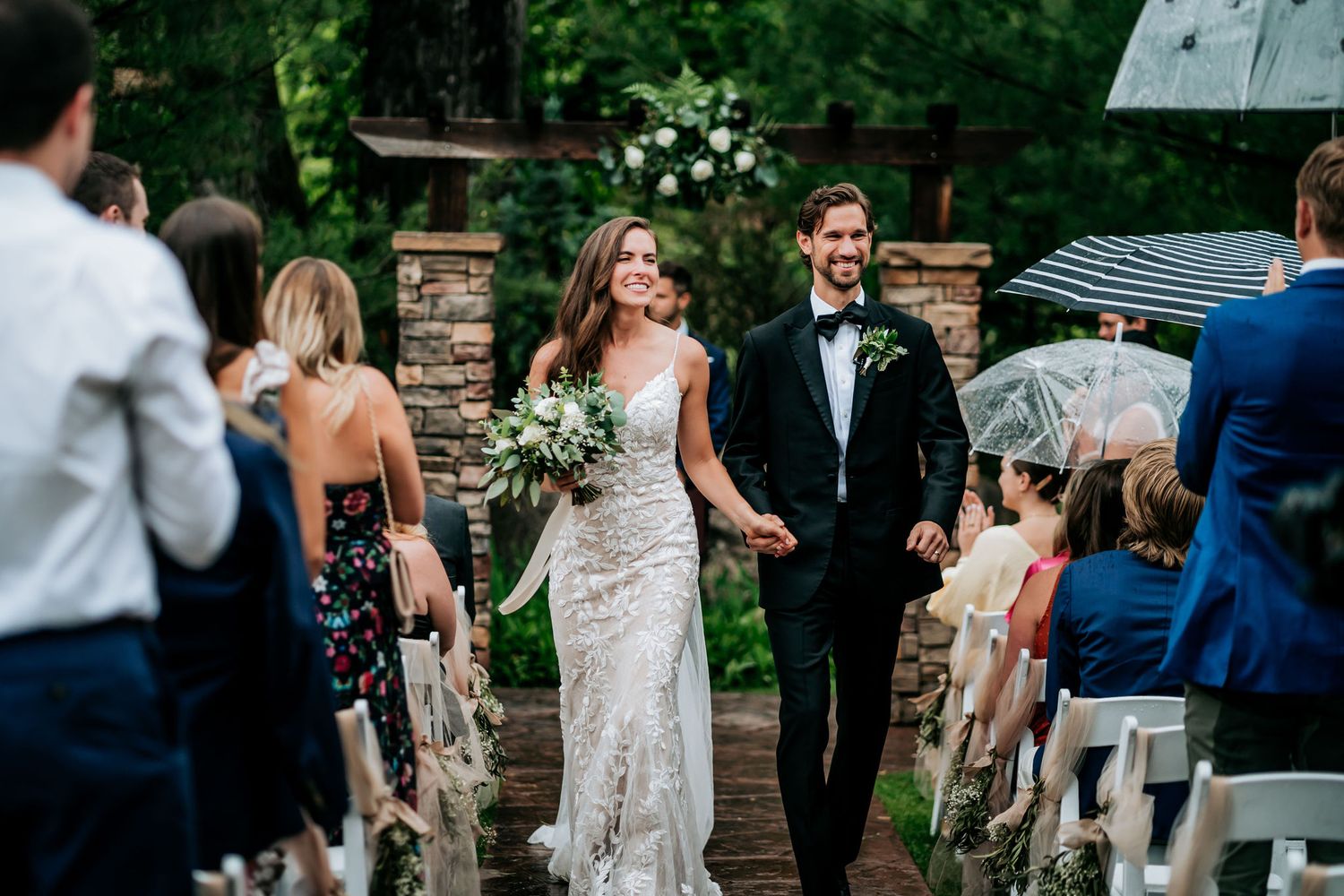 A couple walks down the aisle at an outdoor garden ceremony while guests look on from white chairs.