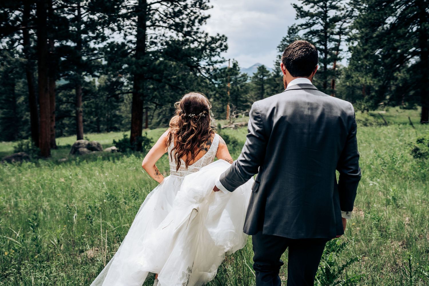A couple walks through a scenic mountain meadow surrounded by evergreen trees on their wedding day.