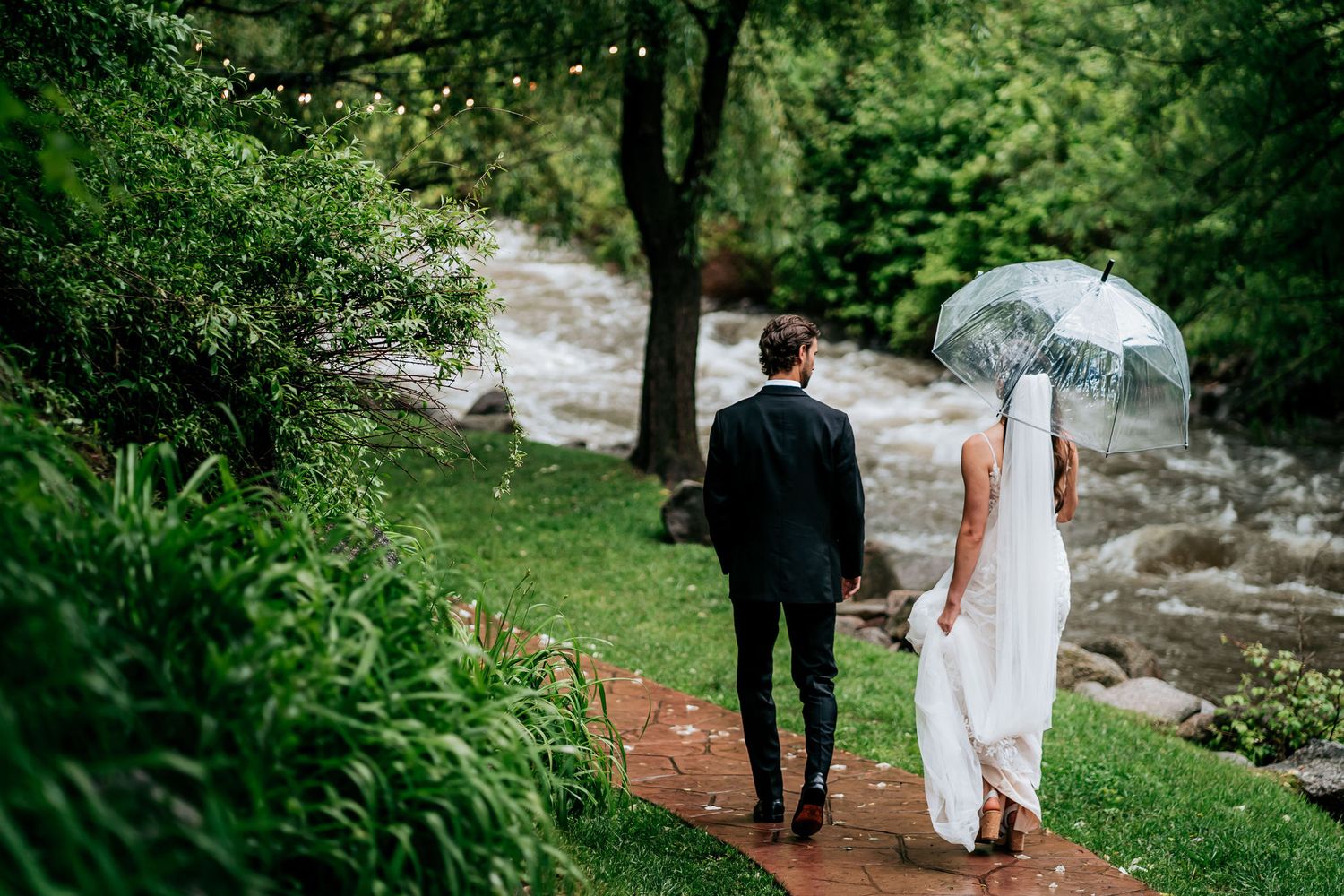 A couple walks along a wooded path by a flowing stream while holding a white umbrella on their wedding day.