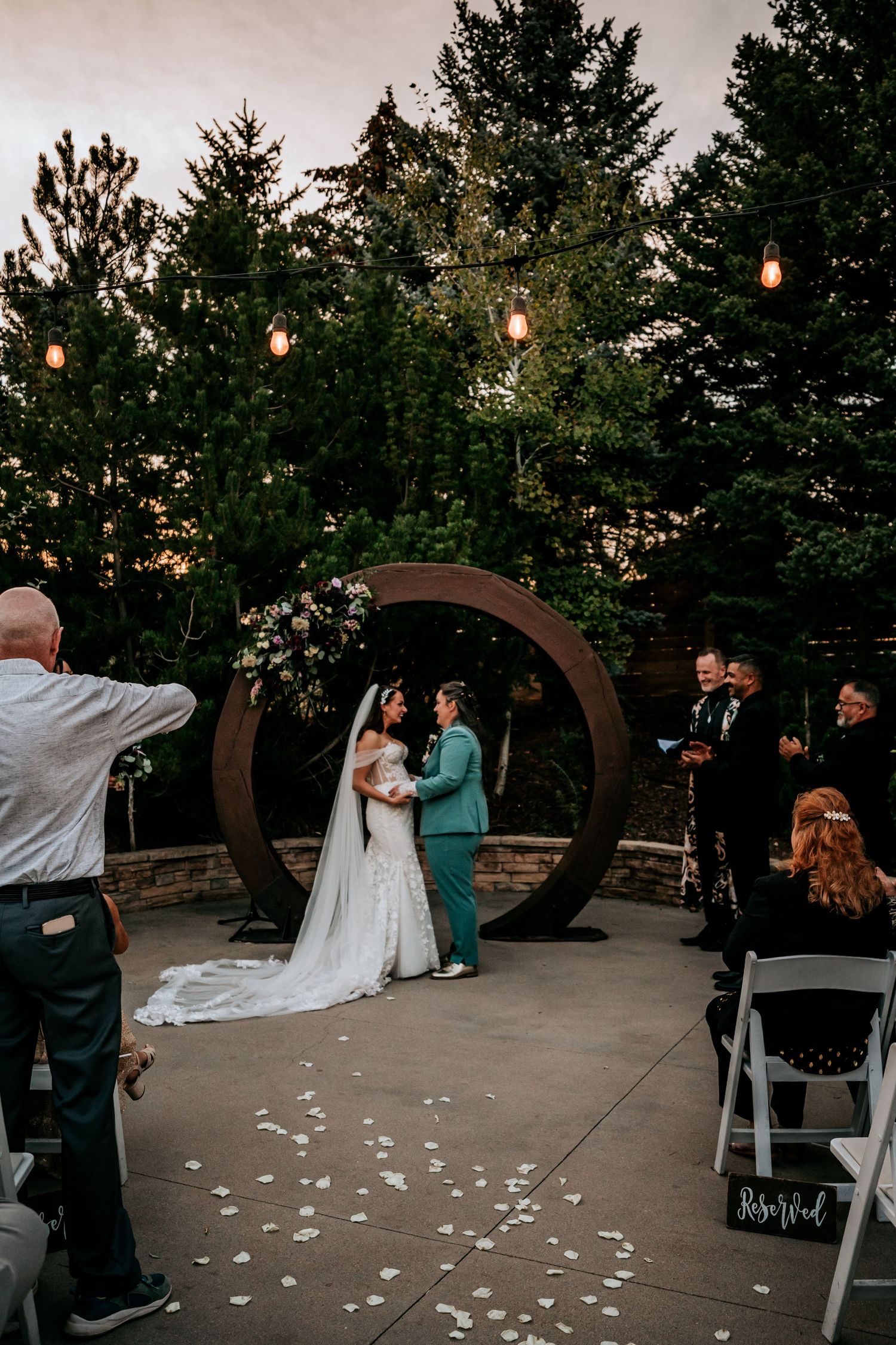 Wide shot of lesbian wedding ceremony at dusk under circular arch with rose petal aisle and string lights.