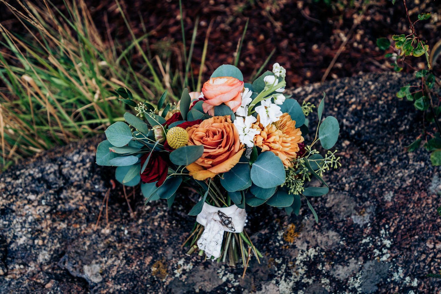 A rustic bridal bouquet with orange roses, eucalyptus leaves, and mixed flowers rests on stone.