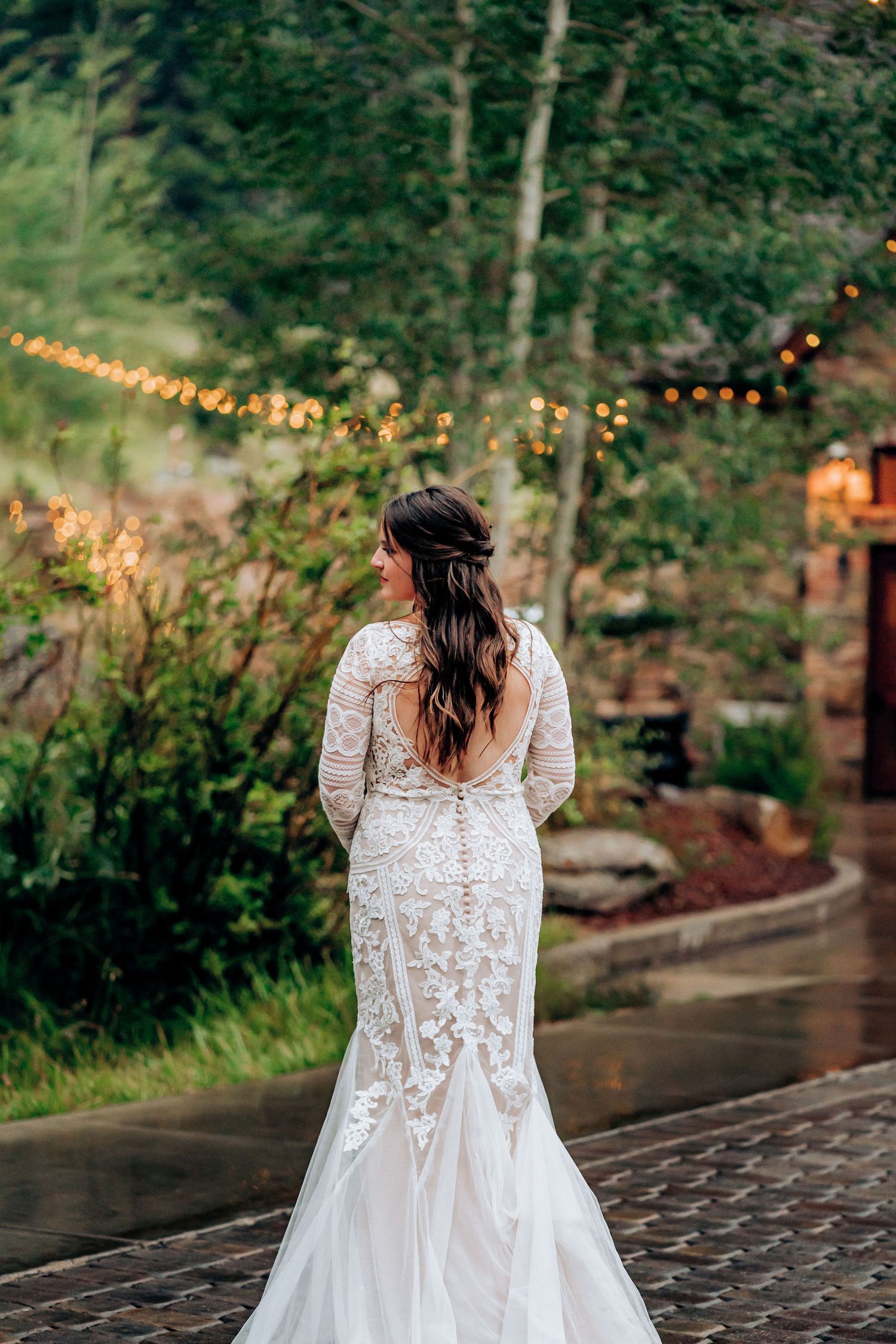 Elegant lace wedding dress with flowing train captured from behind amidst garden string lights.