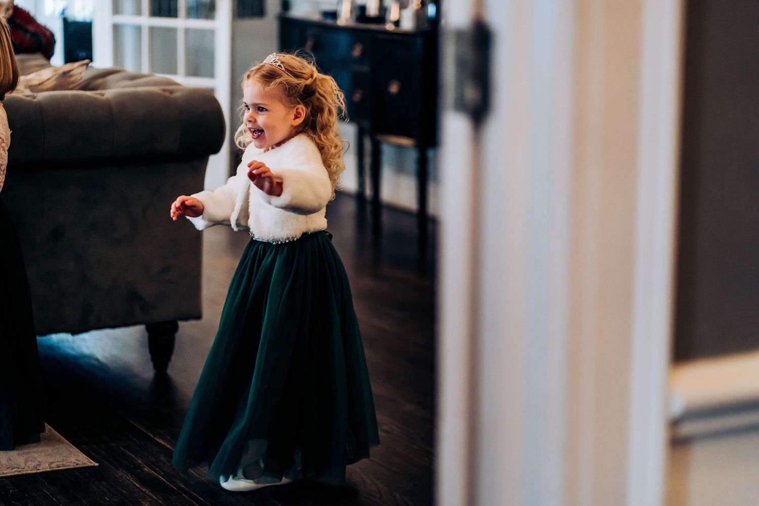 Small child in emerald tulle dress and white fur shrug dancing at wedding reception.