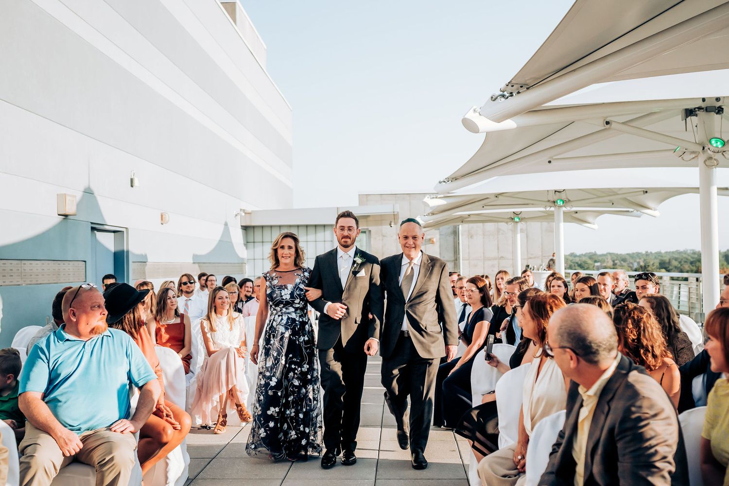 Wedding party members walk down an aisle on a rooftop terrace with guests seated on either side.