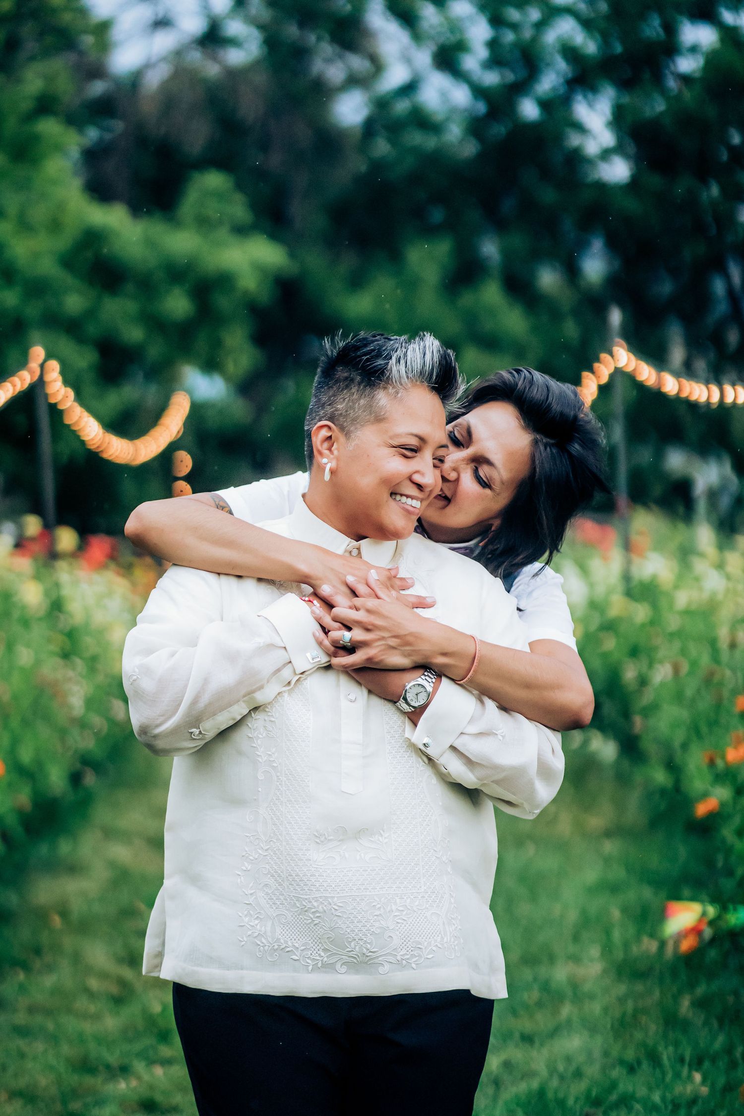 A tender embrace between two people in a garden with string lights and colorful flowers.