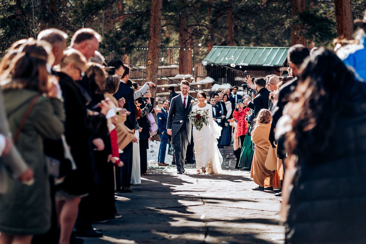 Newlyweds walk down pathway between rows of wedding guests in forest setting.