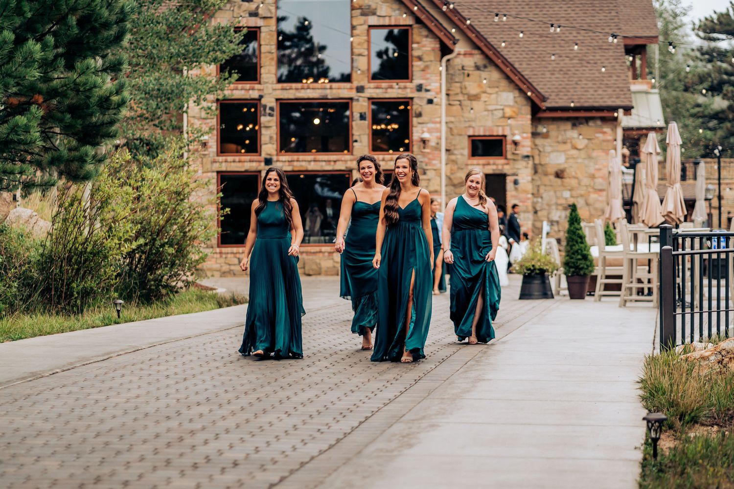 Bridesmaids in teal dresses walk along a path in front of a rustic stone building.