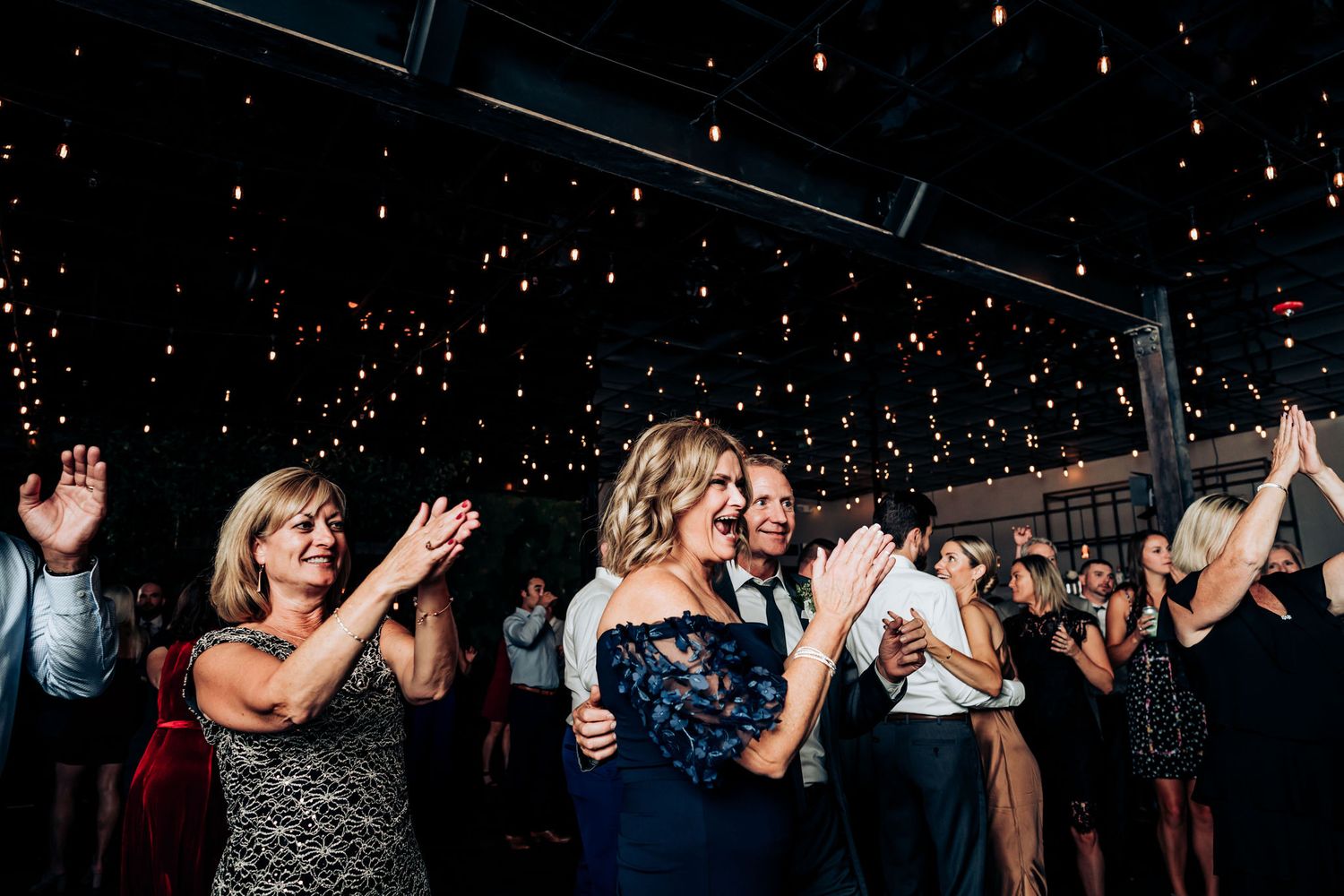 Celebration crowd clapping and dancing under a ceiling filled with twinkling fairy lights.