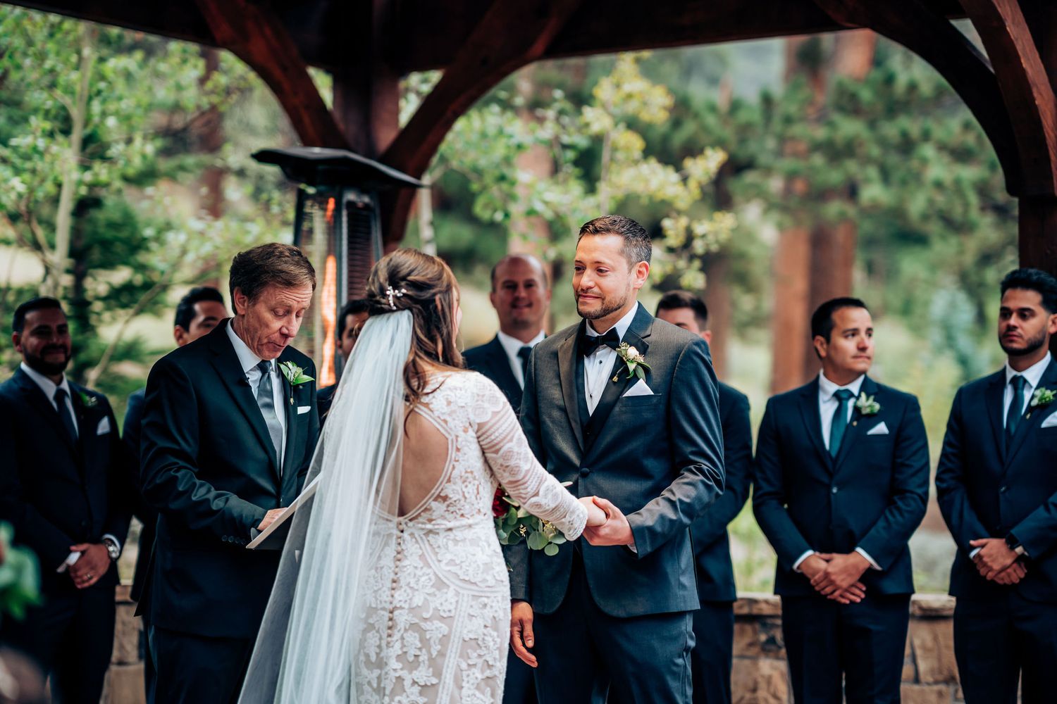 A wedding ceremony takes place under a wooden pavilion surrounded by lush greenery and trees.