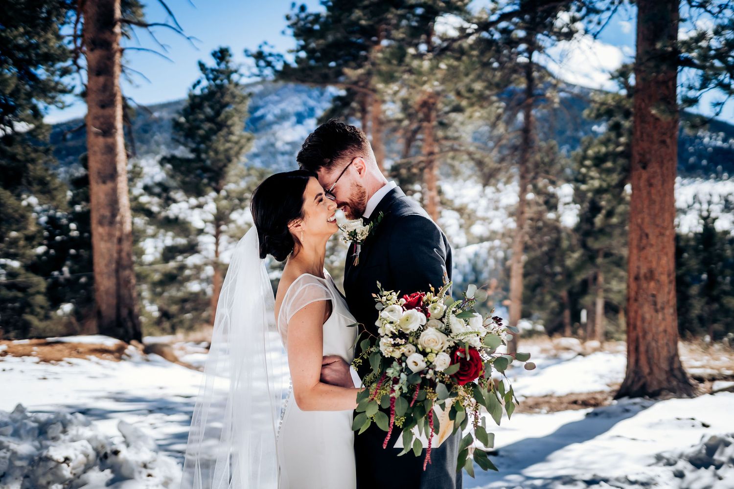 Newlyweds share an intimate moment surrounded by snow-covered pine trees in a winter mountain landscape.