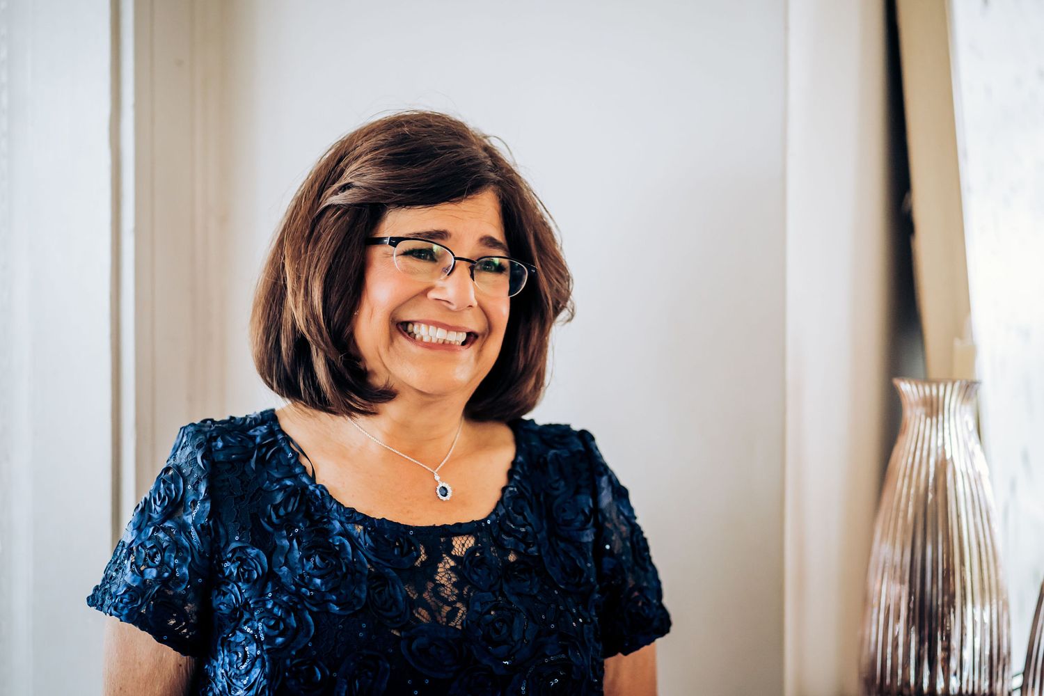 A woman wearing a navy blue lace dress smiles warmly in an indoor setting.