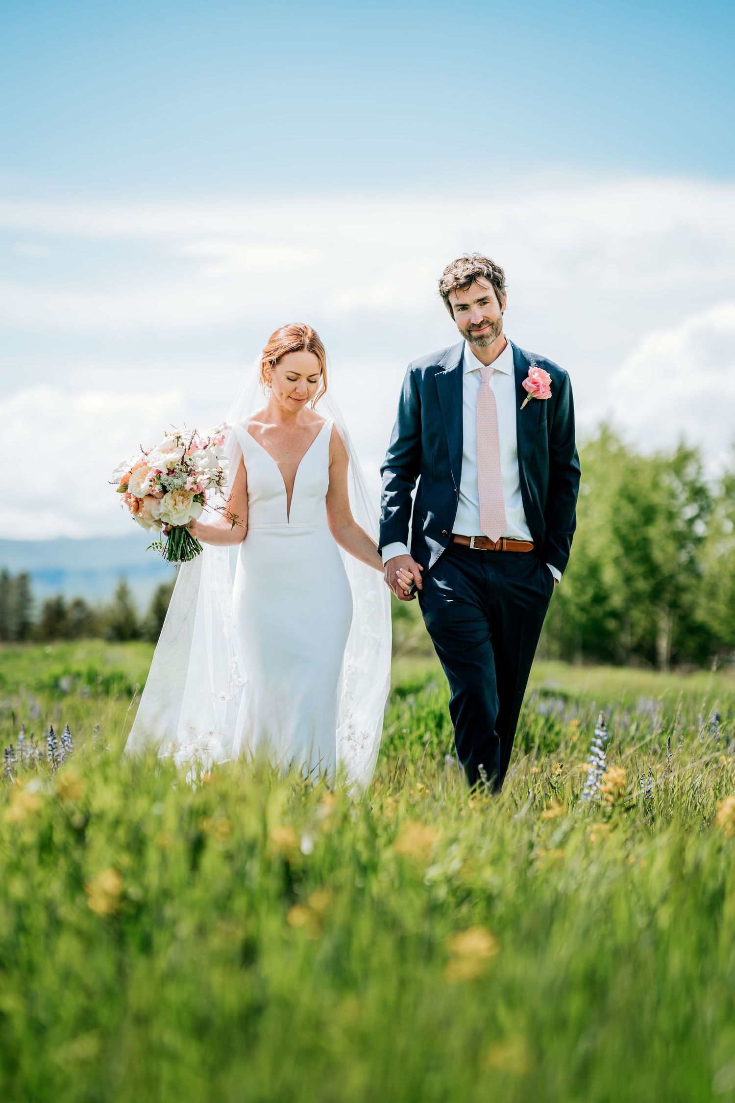 Newlyweds walk hand in hand through a lush meadow with mountains and blue sky in the background.