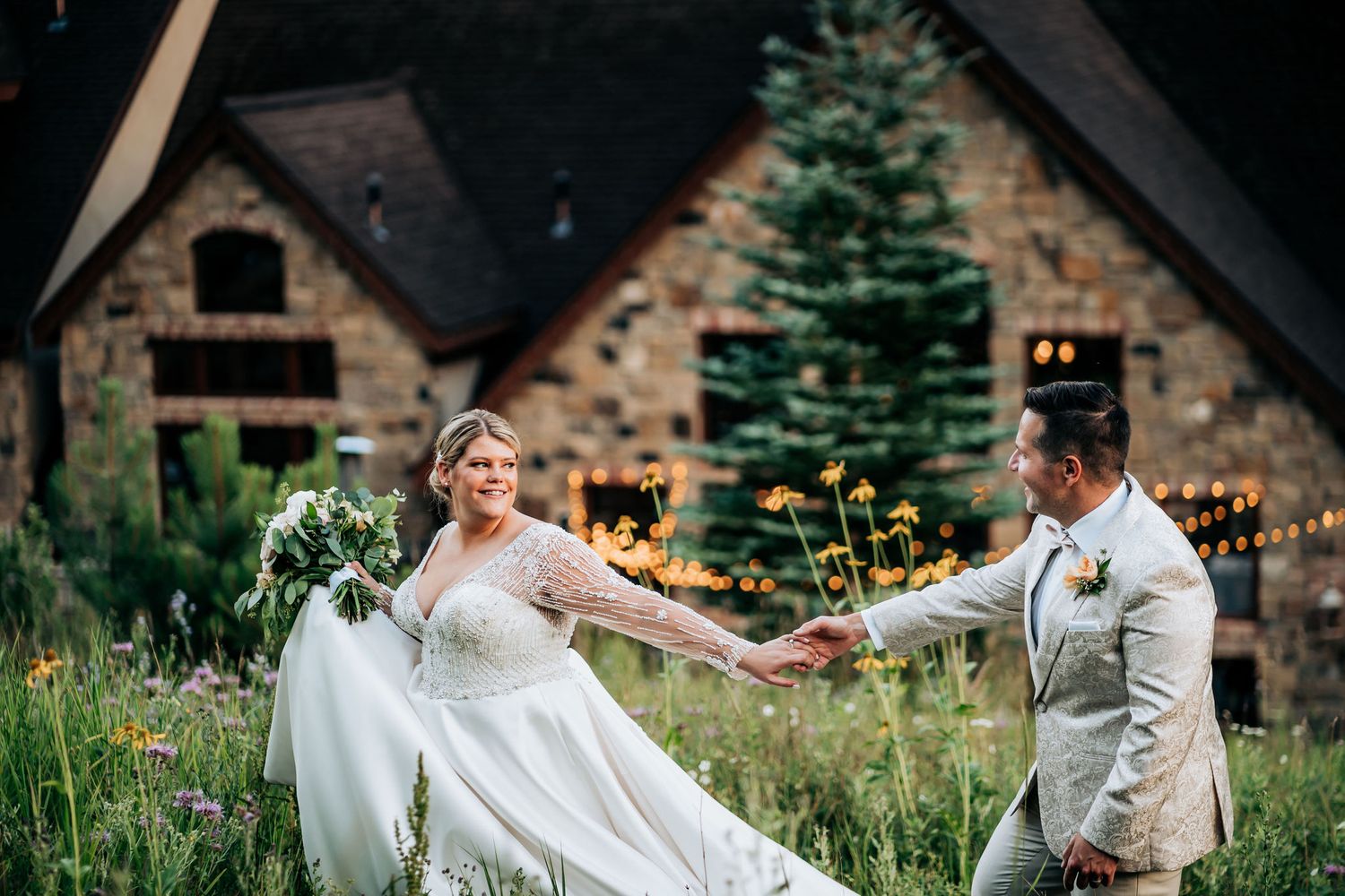 Couple shares playful moment outside rustic stone lodge surrounded by wildflowers.