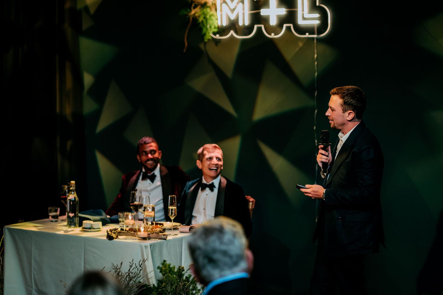 Three people seated at a table during a comedy show event with geometric backdrop and M+L signage.