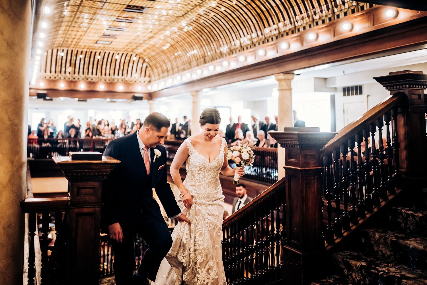 Elegant couple descends ornate wooden staircase at historic venue during wedding celebration.