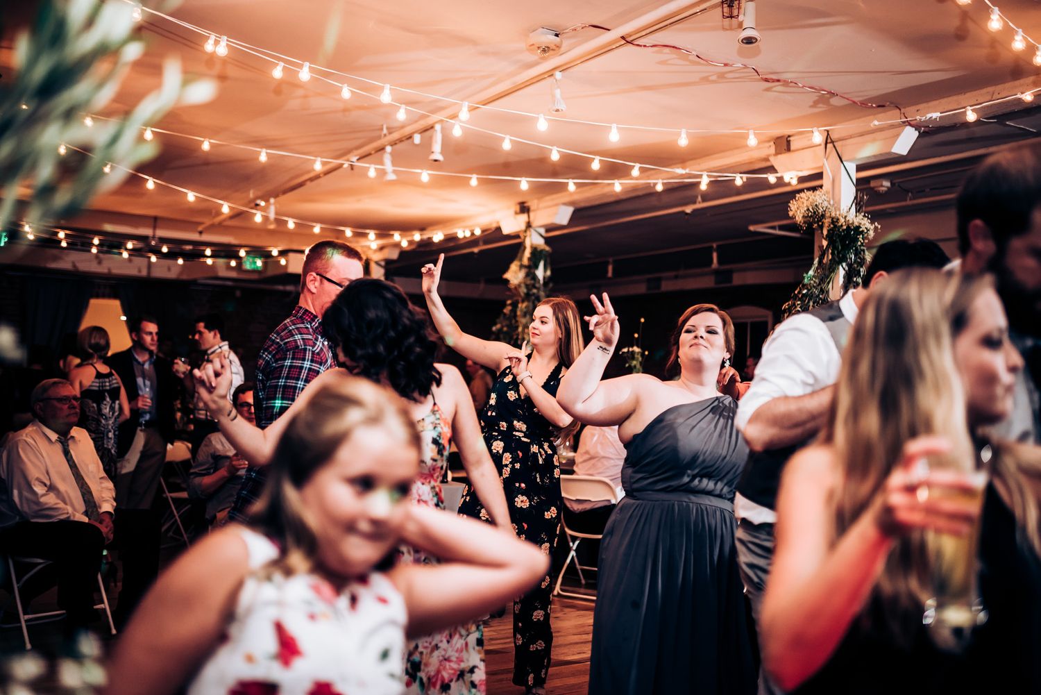 A crowded dance floor filled with celebratory guests dancing under warm string lights at a wedding reception.