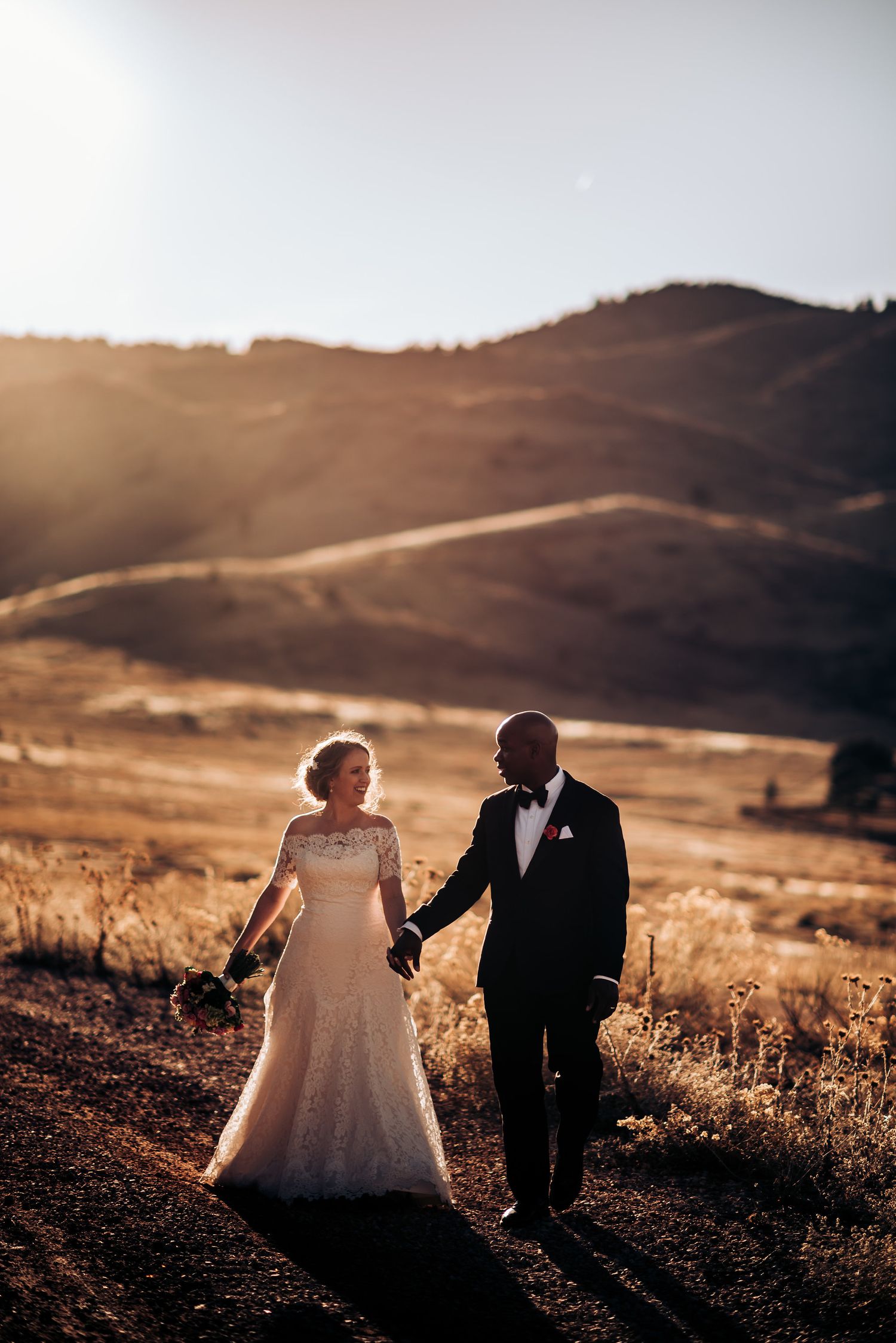 Newlyweds hold hands while walking along a scenic mountainside trail at golden hour.