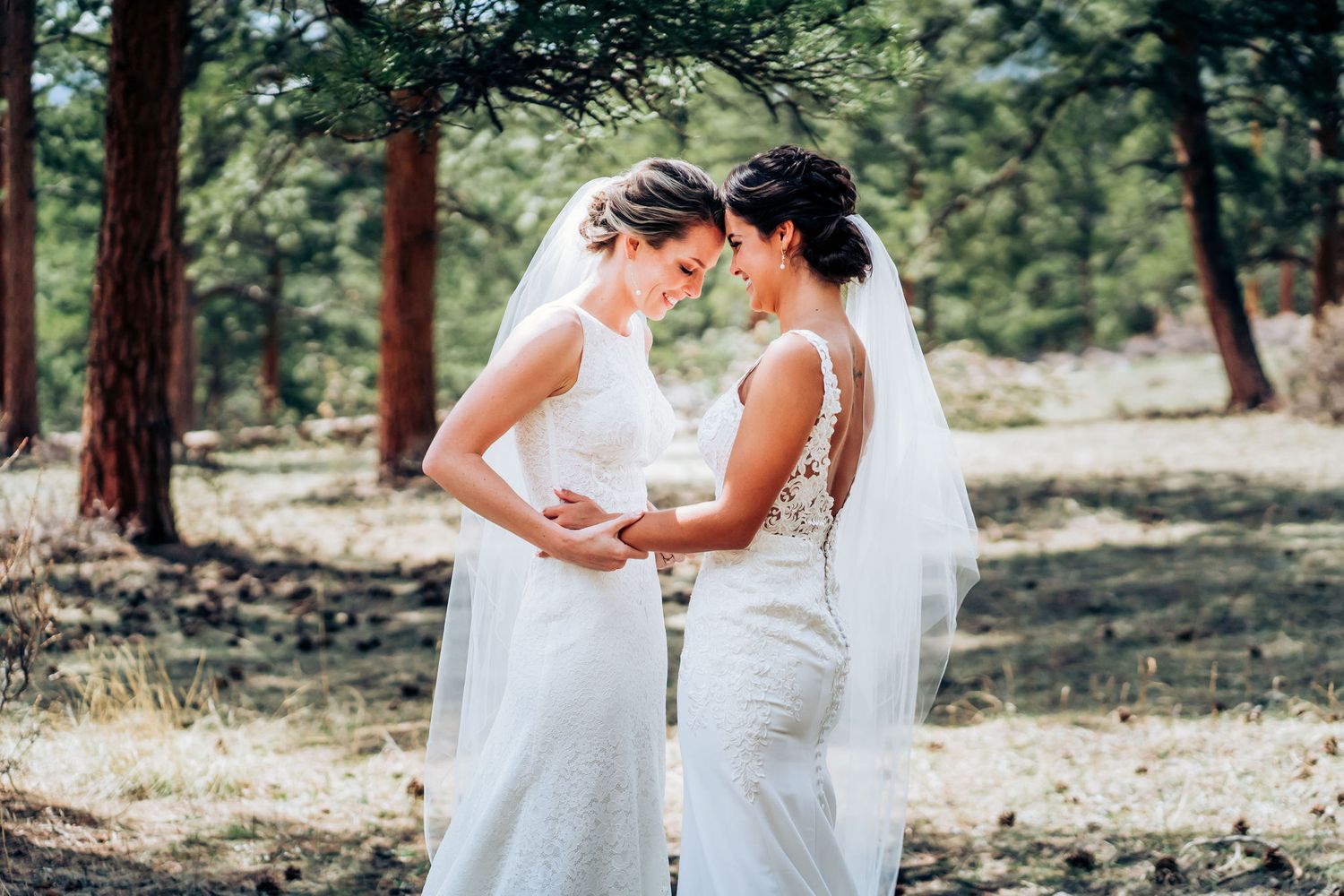 Two brides share a close moment in white wedding gowns surrounded by pine trees.