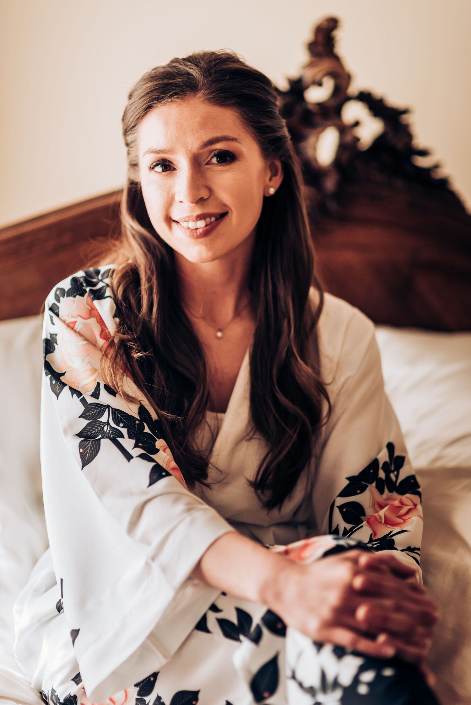 A woman in a floral white robe sits on an elegant bed during wedding preparations.