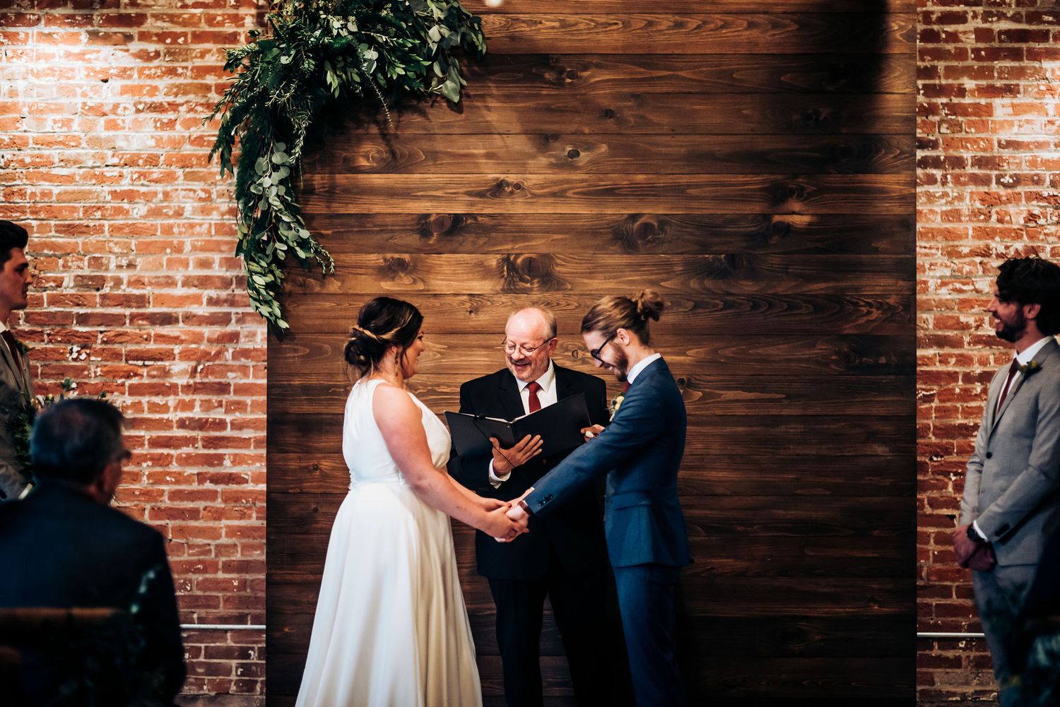 Two people exchange wedding vows in an intimate ceremony against a rustic wooden wall with exposed brick backdrop.