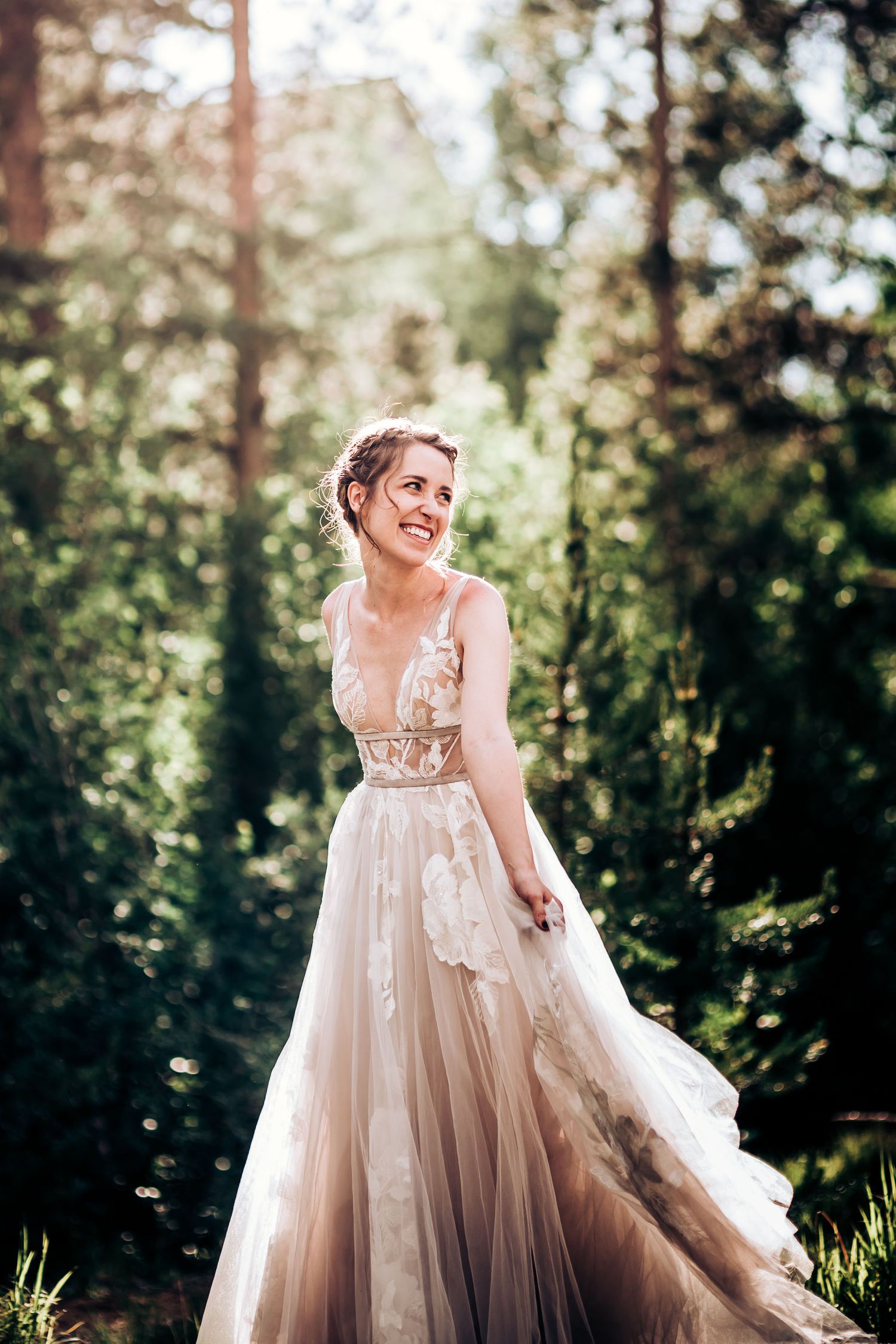 A joyful bride in a blush tulle gown laughs while standing in a sunlit forest clearing.