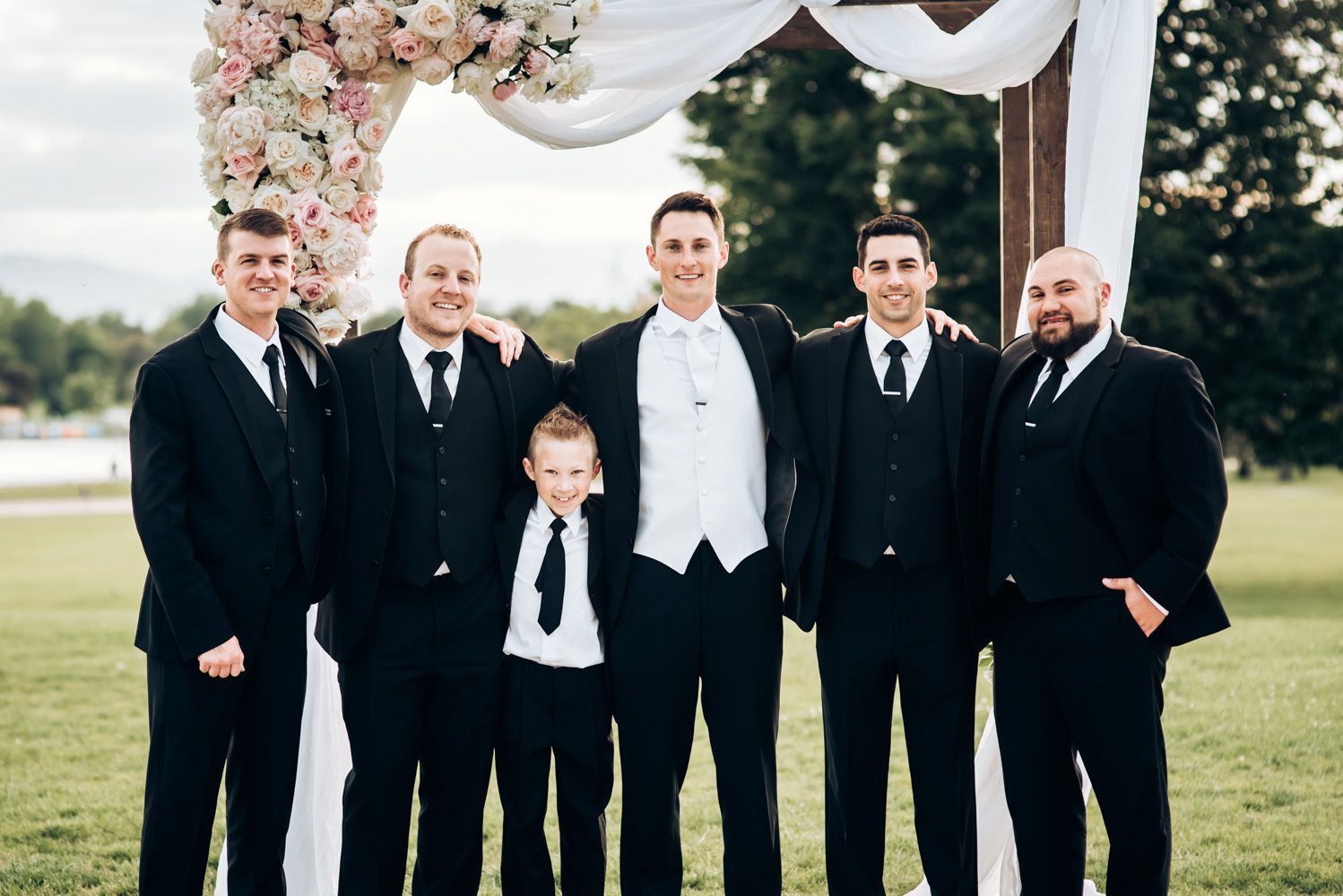 Groomsmen and young ring bearer in formal attire pose together on sunny lawn.