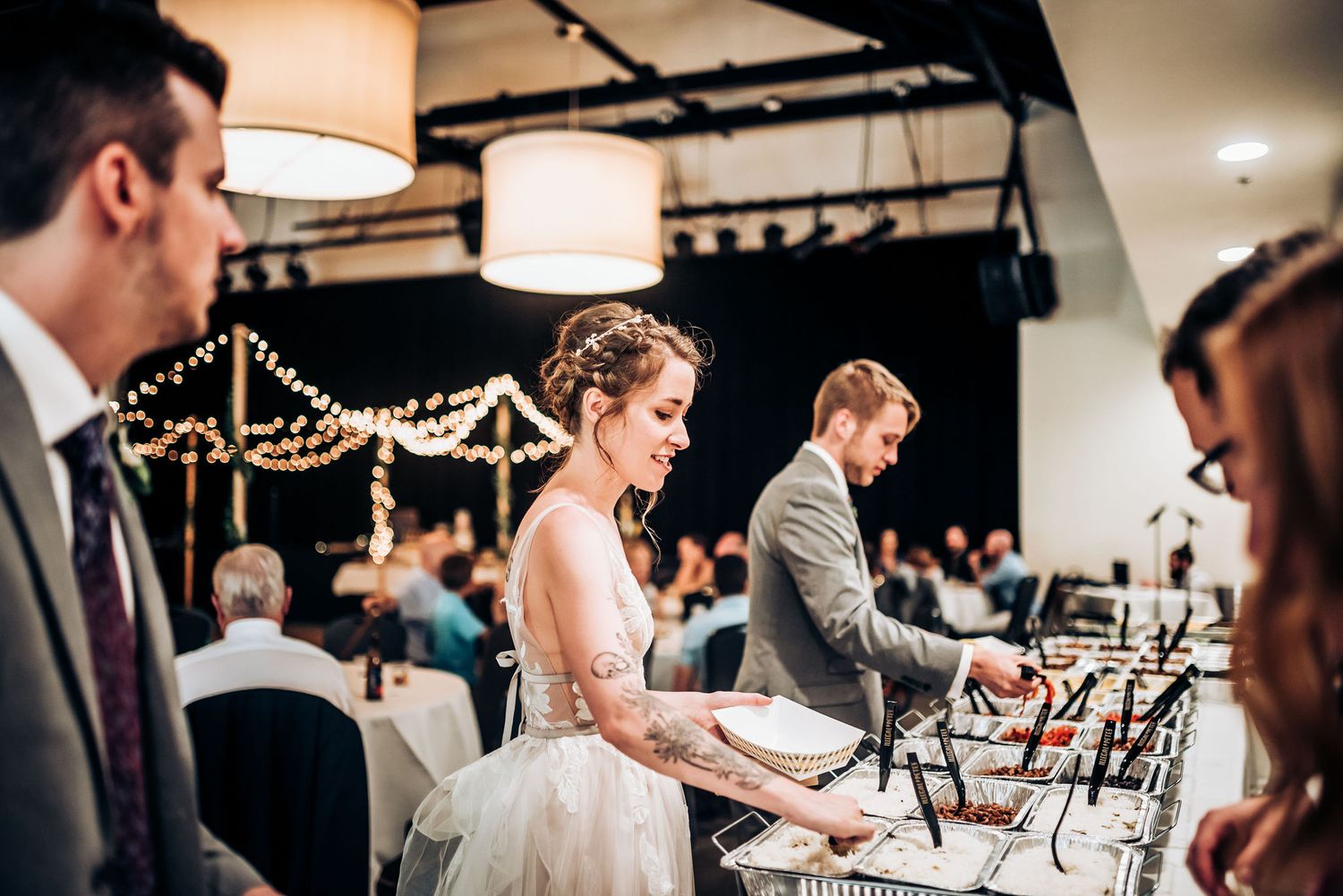 Wedding reception buffet line with guests serving food under decorative string lights.