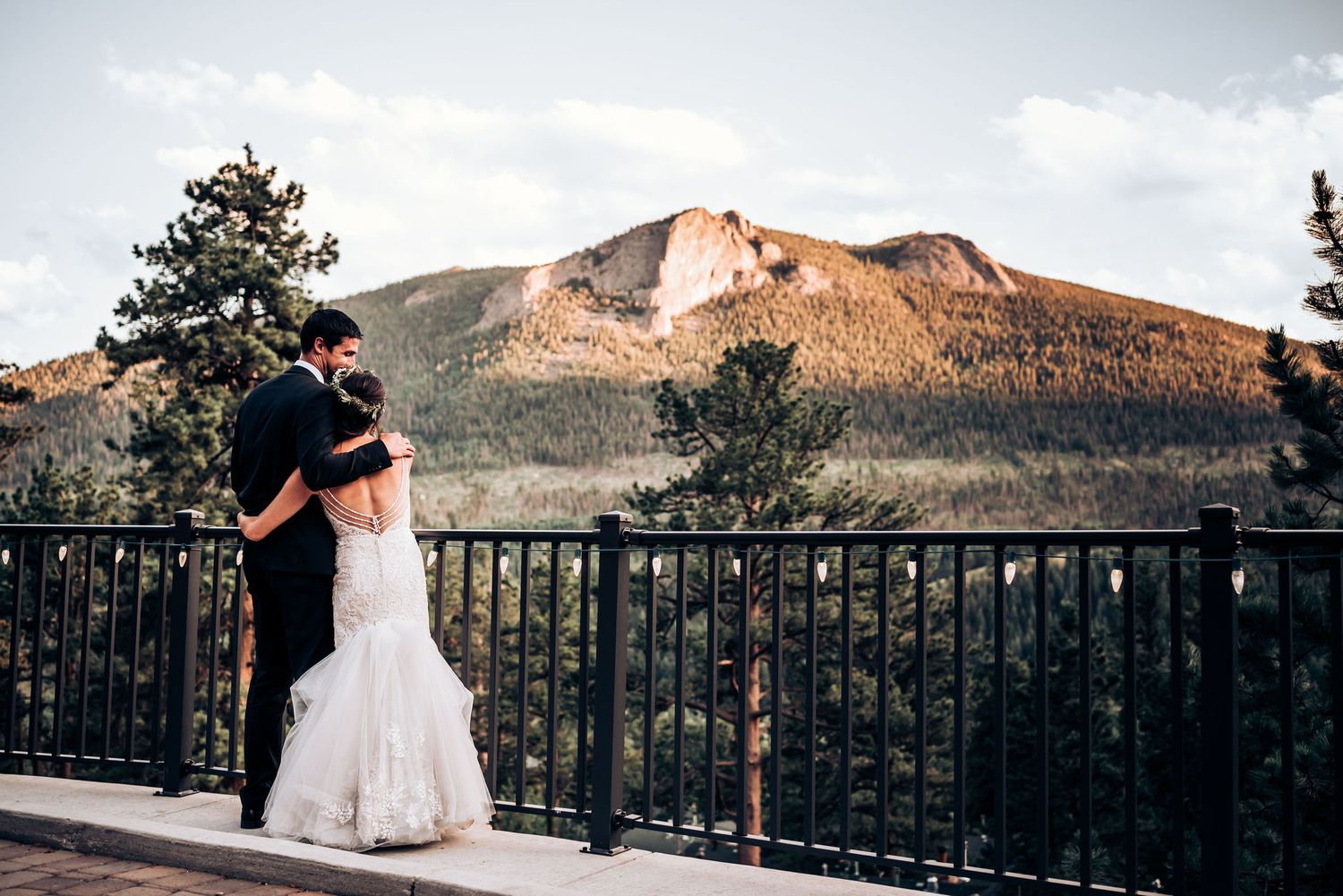Newlyweds embrace on balcony overlooking stunning mountain vista at sunset.